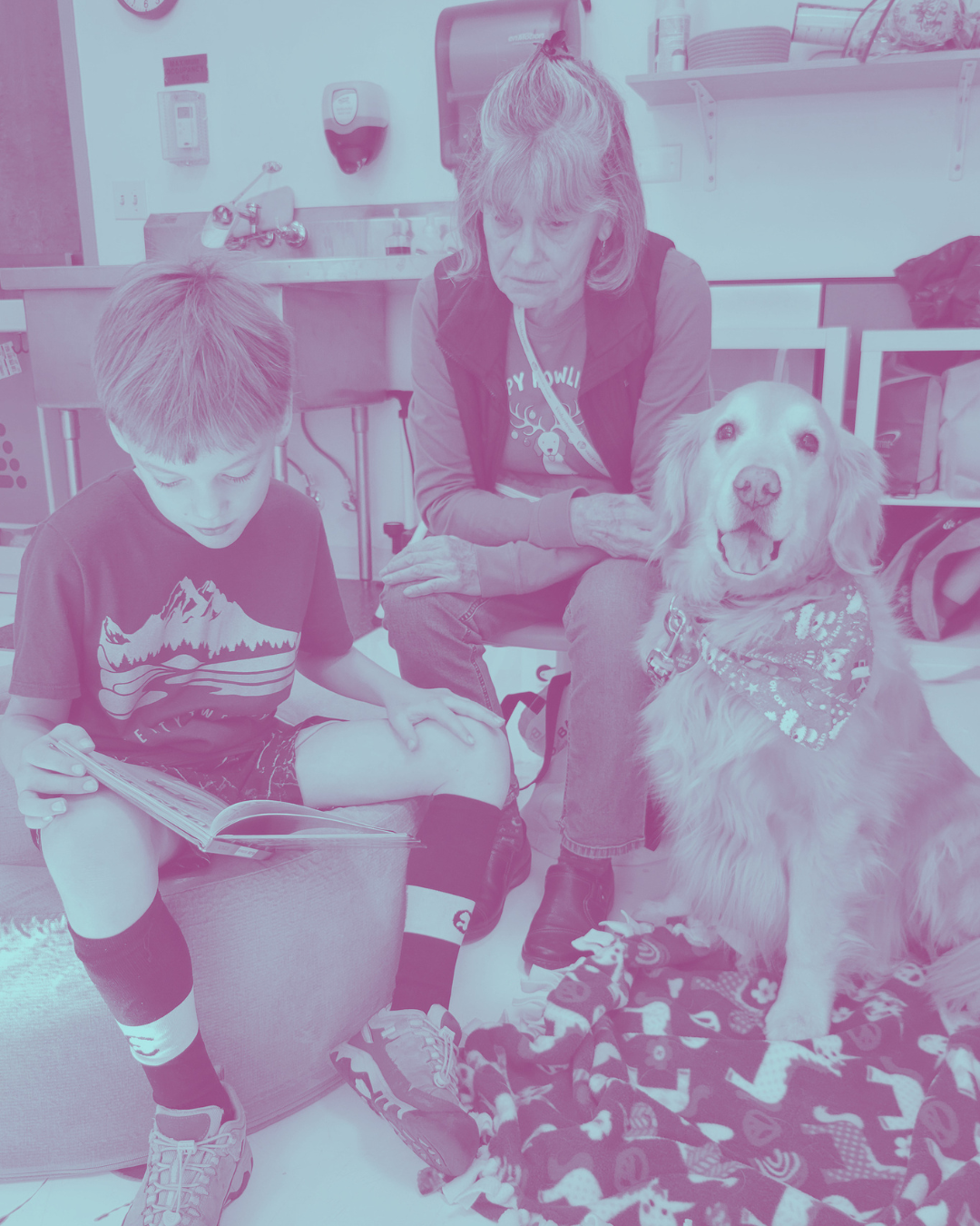 A woman, a boy, and a dog sitting in a veterinary clinic. The boy is reading a book, and the dog is wearing a bandana and sitting on a blanket. The woman is observing the dog.