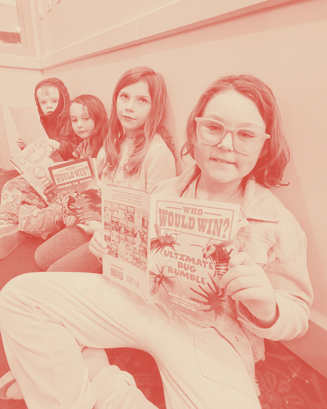 Four children sitting on a bench, holding comic books and reading, against a wall.