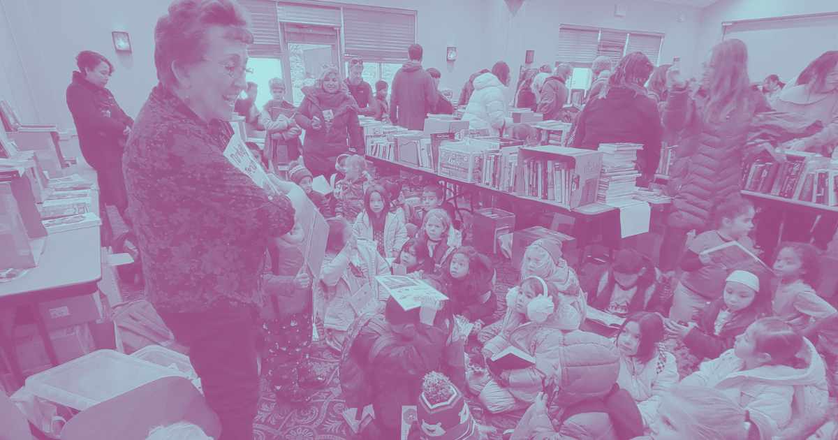 A crowded indoor book fair with children sitting on the floor and adults browsing books displayed on tables.