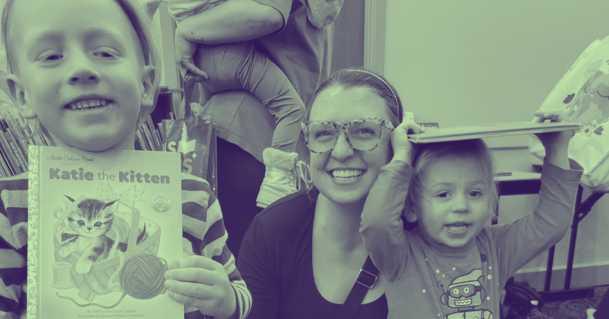 A woman with glasses and a headband smiling at the camera, flanked by two young girls. One girl is holding a children's book titled 'Katie the Kitten' and the other girl is holding an object above her head. The background shows a room with books and 