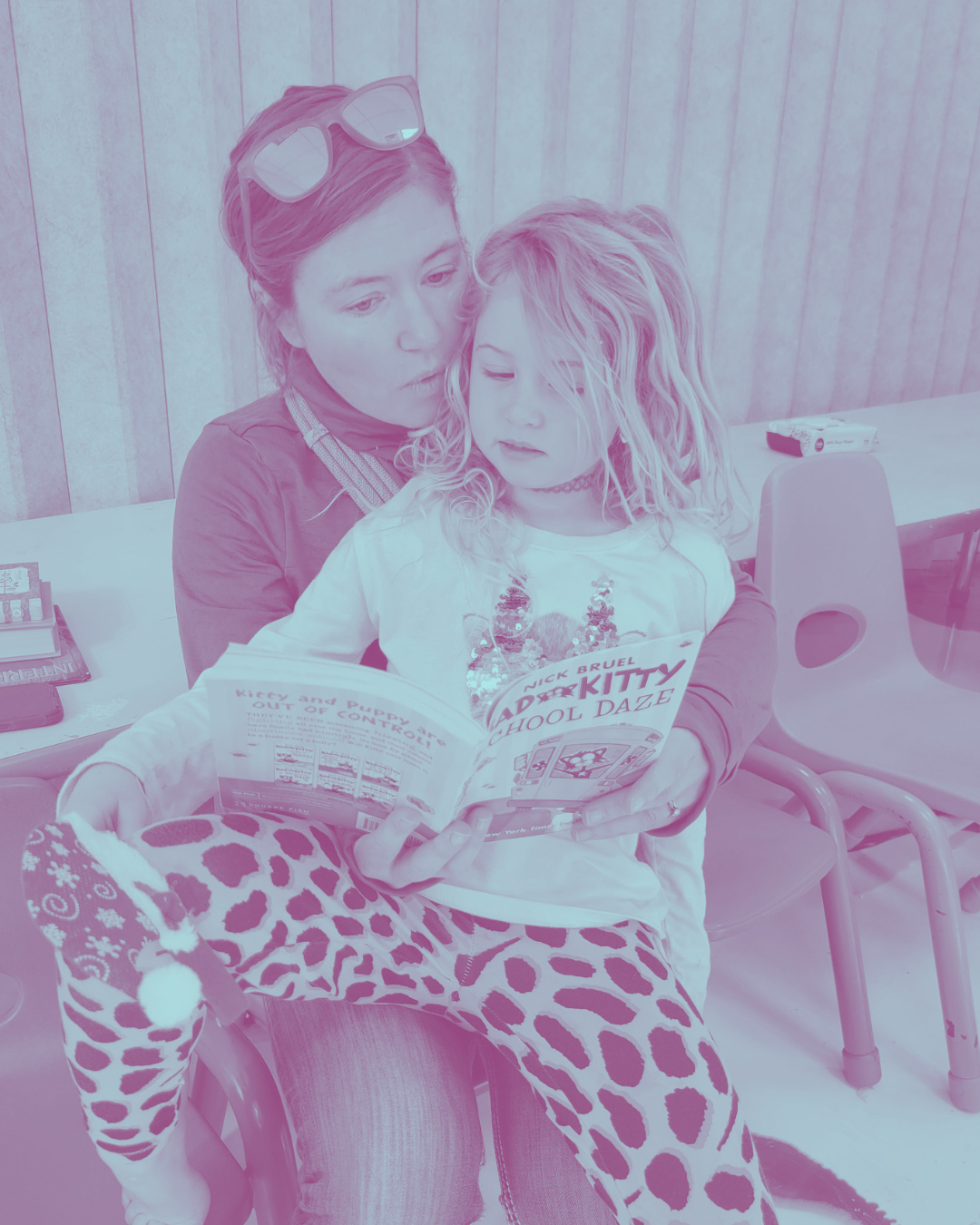 A woman and a young girl sitting together, reading a comic book titled 'Adi Kitty School Daze'. The girl is wearing leopard-print pajamas and has curly hair, while the woman has red hair and sunglasses on her head.