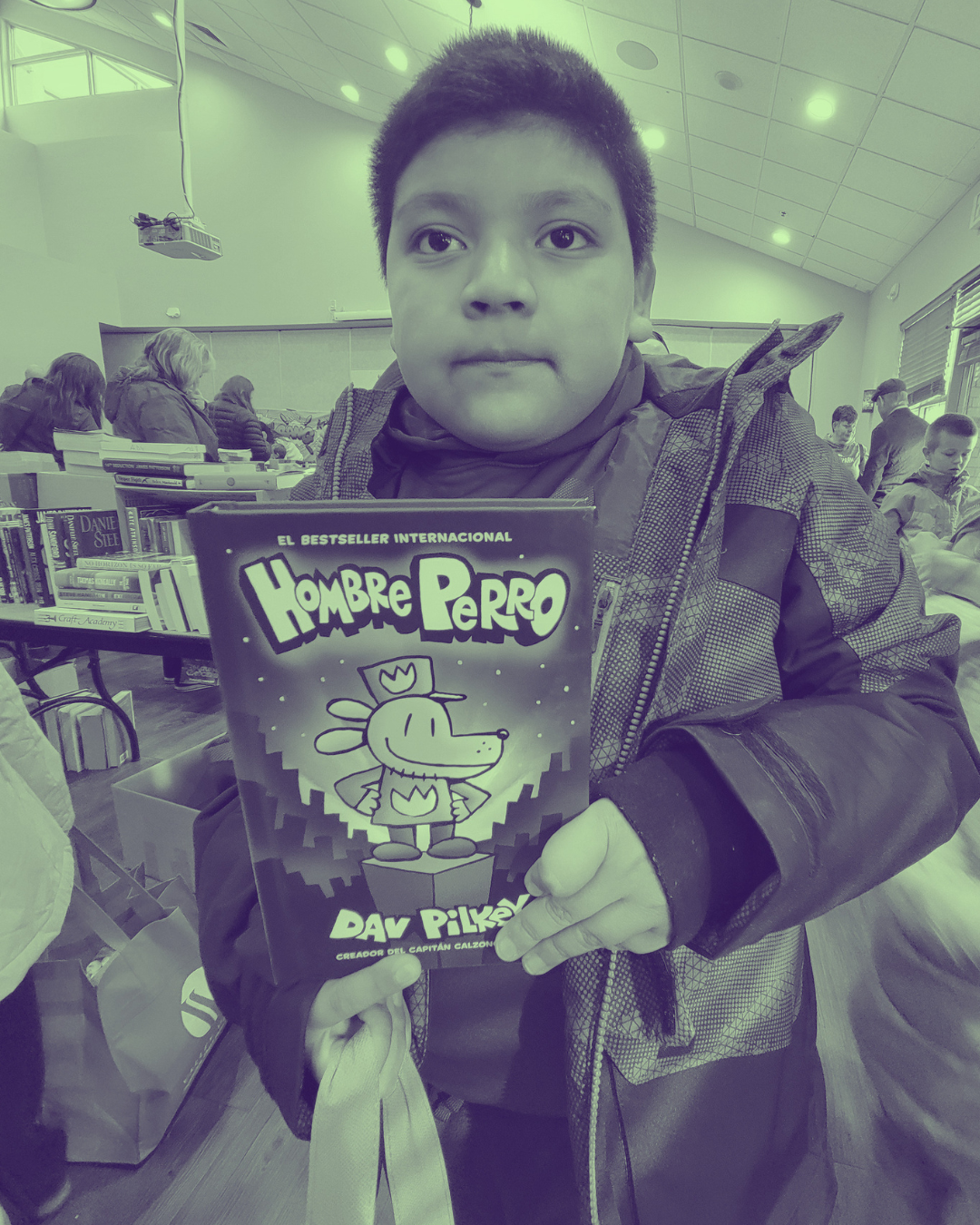 A boy holding a book titled "Hombre Perro" in a library or bookstore, with other people browsing bookshelves in the background.