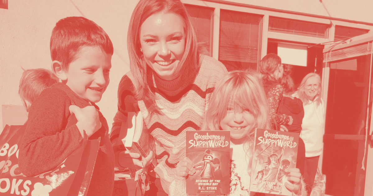 Smiling woman and two children holding books outside a bookstore, with other people in the background.