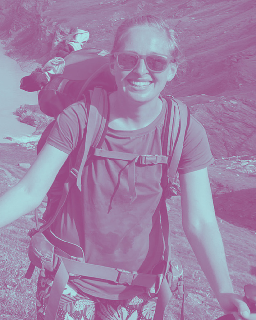 A woman hiking outdoors with a backpack, wearing sunglasses, a t-shirt, and patterned shorts, smiling at the camera.