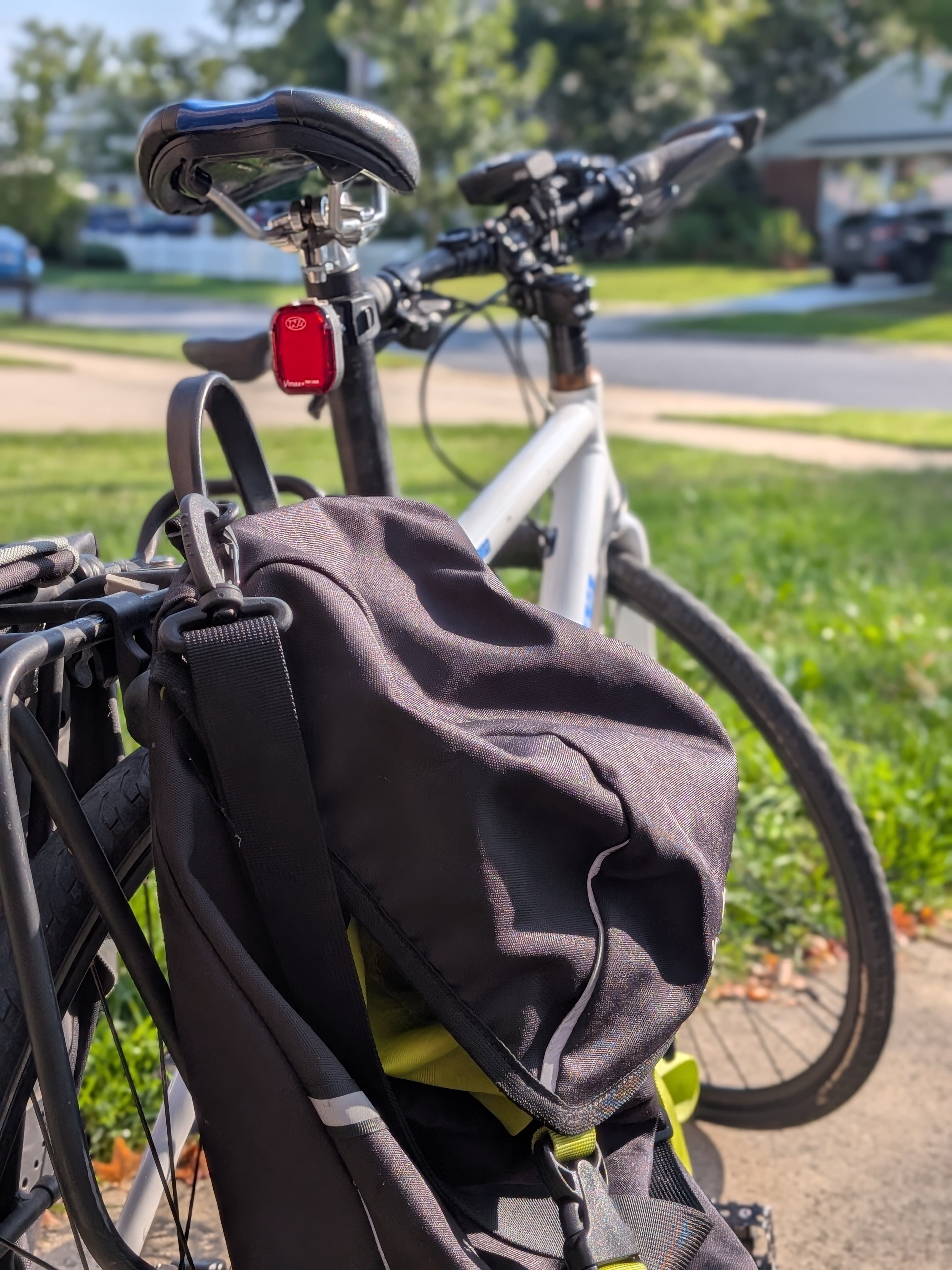 Close-up of a bicycle seat and a black backpack attached to a bicycle frame outdoors on a sunny day, with trees and parked cars in the background.