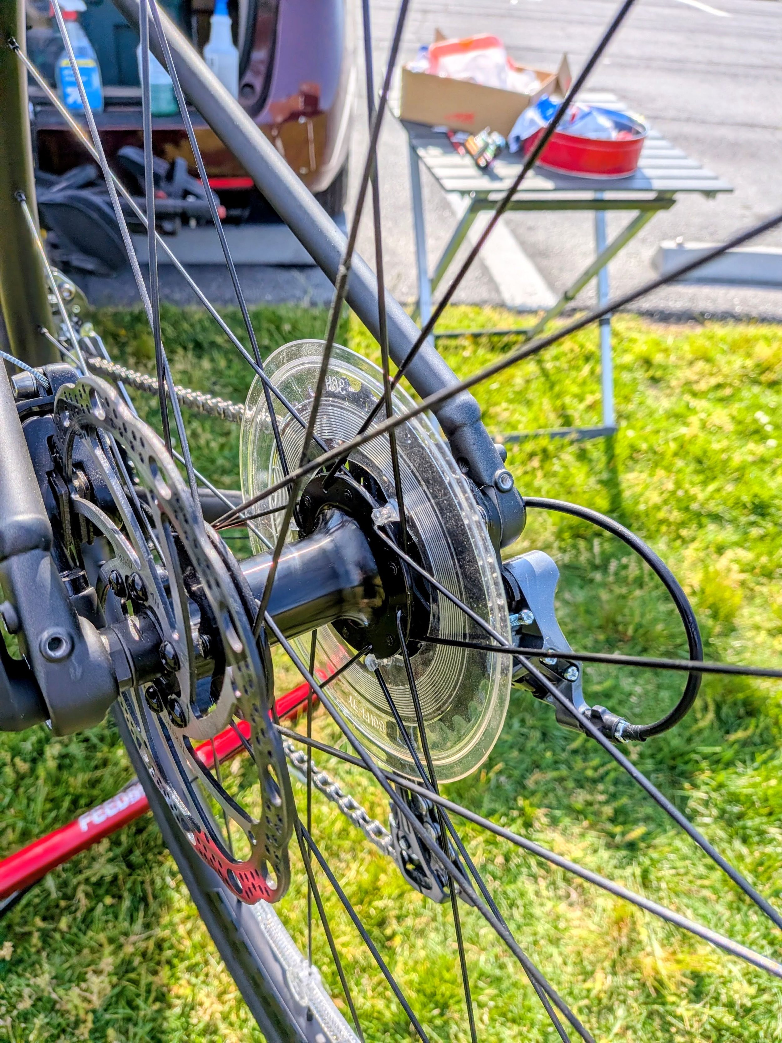 Close-up of a bicycle wheel with disc brake rotor, spokes, and hub, with a grassy background and a preparation table with snacks and supplies in the background.