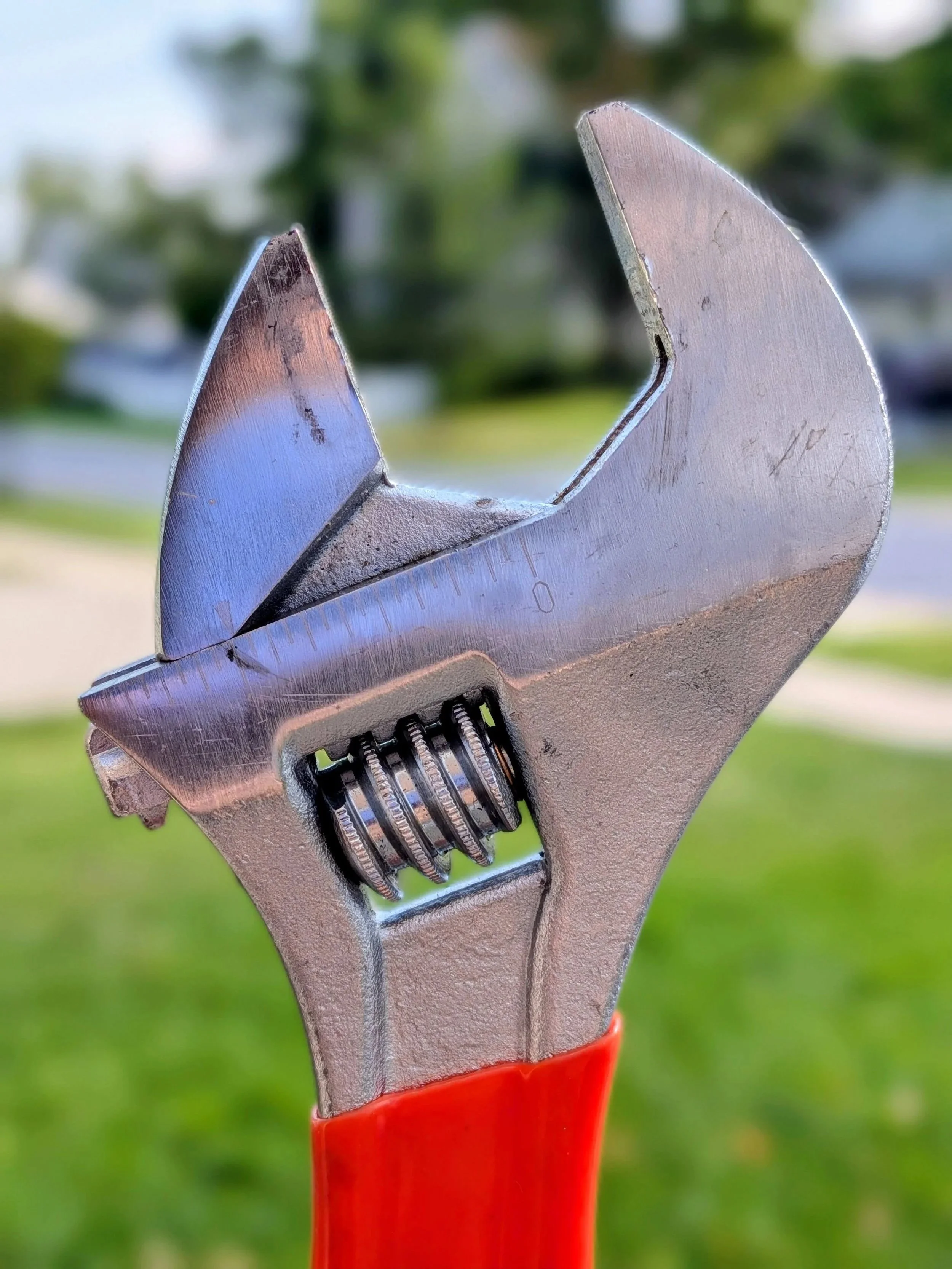 Close-up of an adjustable wrench with a red handle, with the background of green grass and trees.