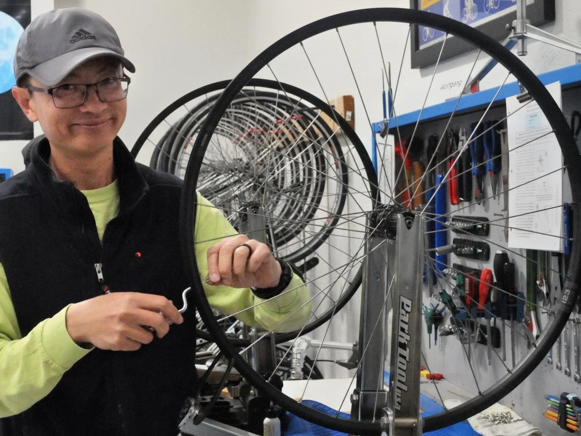 A person wearing glasses and a gray Adidas cap is working on a bicycle wheel in a workshop. The background shows shelves with tools and multiple bicycle wheels lined up.
