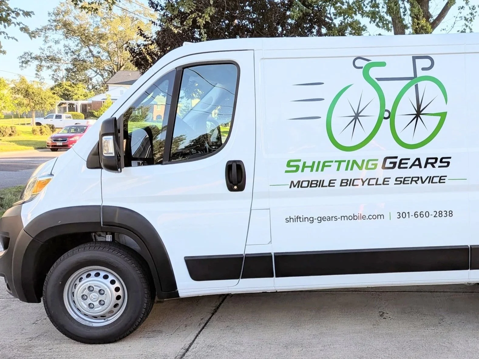 White service van with logo and text for Shifting Gears Mobile Bicycle Service, featuring a green bicycle graphic and contact information, parked on a residential street with trees and houses in the background.