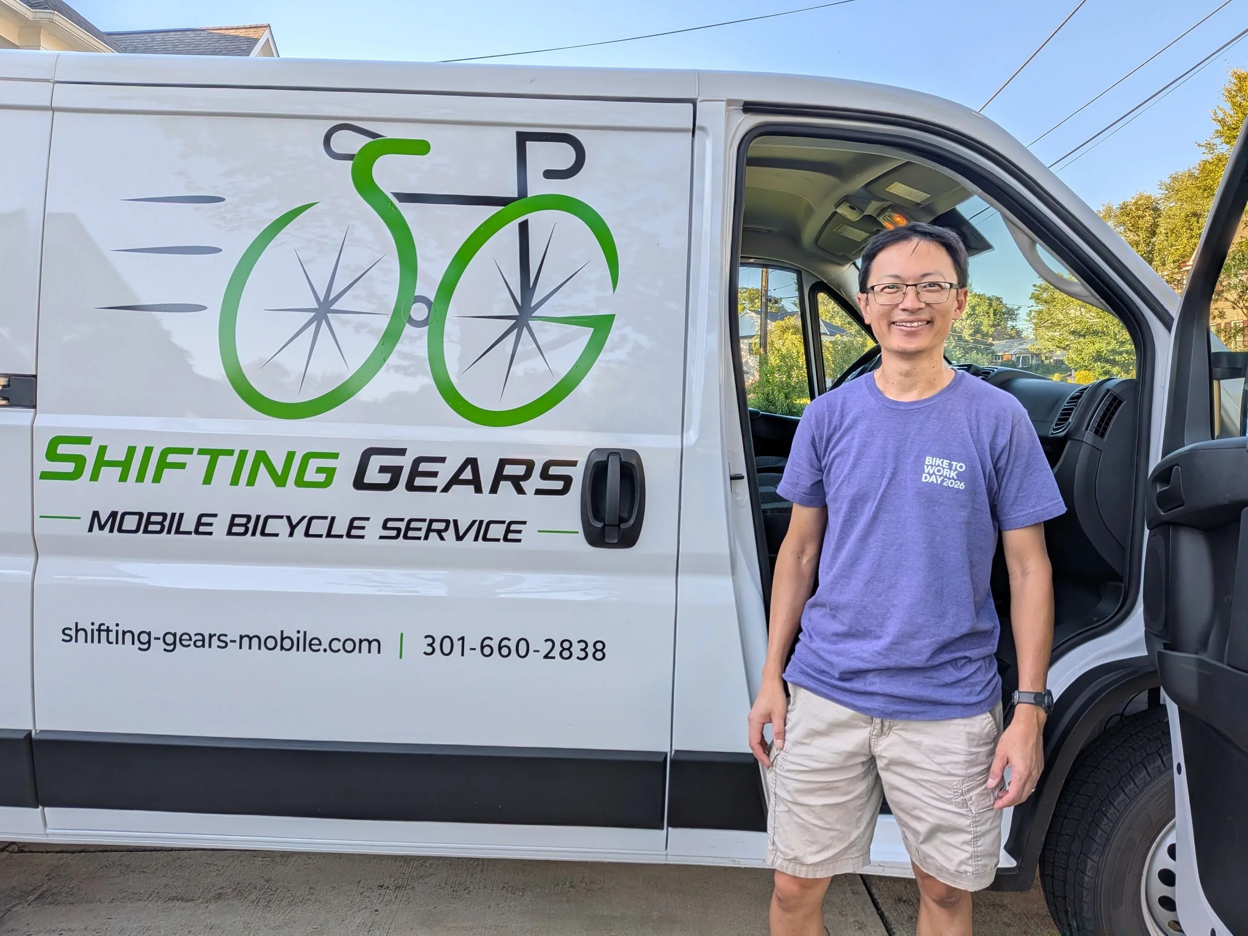A smiling man wearing glasses, a purple T-shirt that says 'Bike to Work Day 2026,' and beige shorts stands next to a white van with a logo of a green bicycle and the words 'Shifting Gears' and 'Mobile Bicycle Service' on the side. The van is parked outdoors on a residential street with trees and houses in the background.