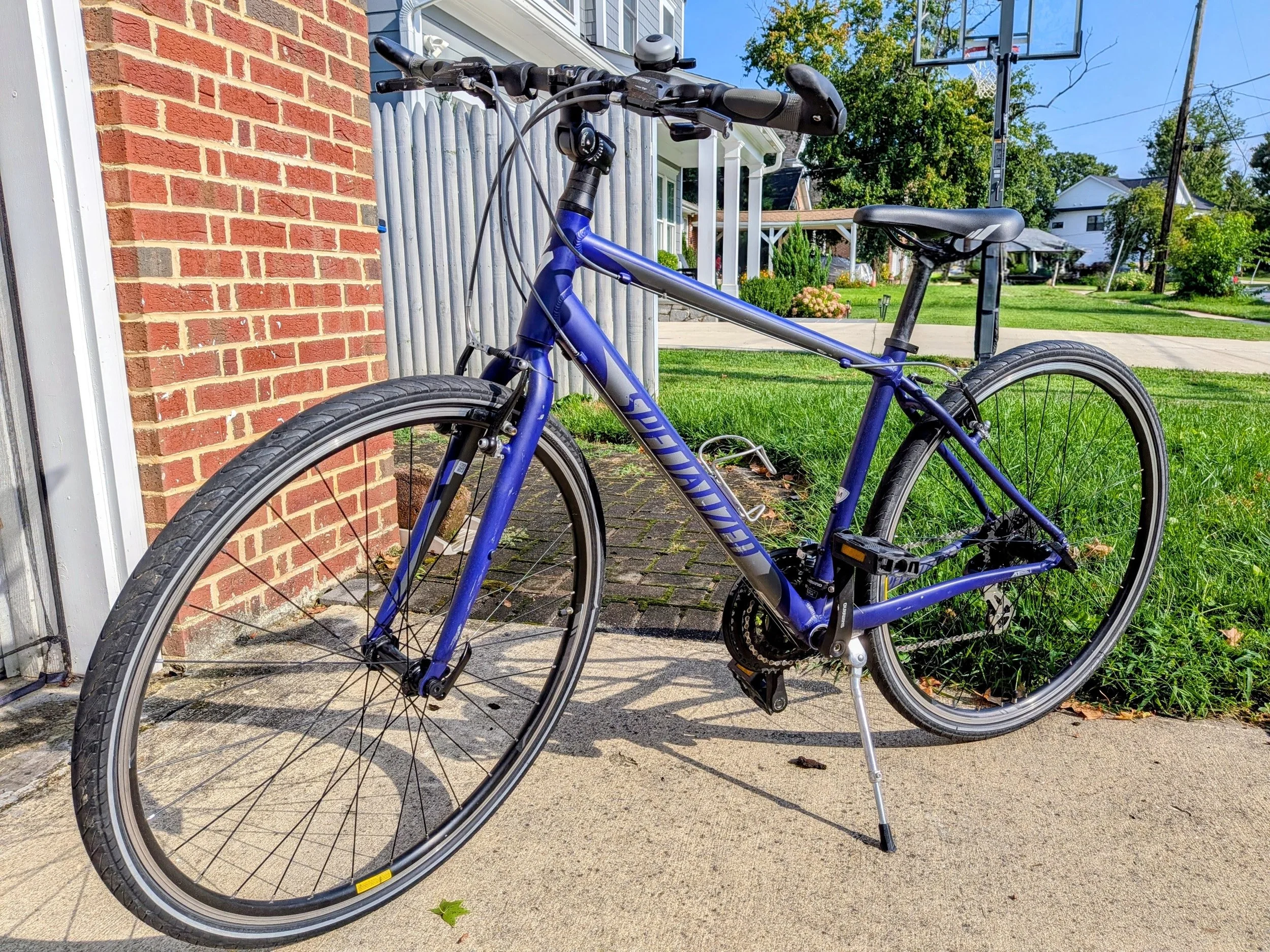 Blue bicycle with black seat and handlebars, standing on a kickstand on a concrete sidewalk near a brick house wall. In the background, there are trees, houses, and a basketball hoop.