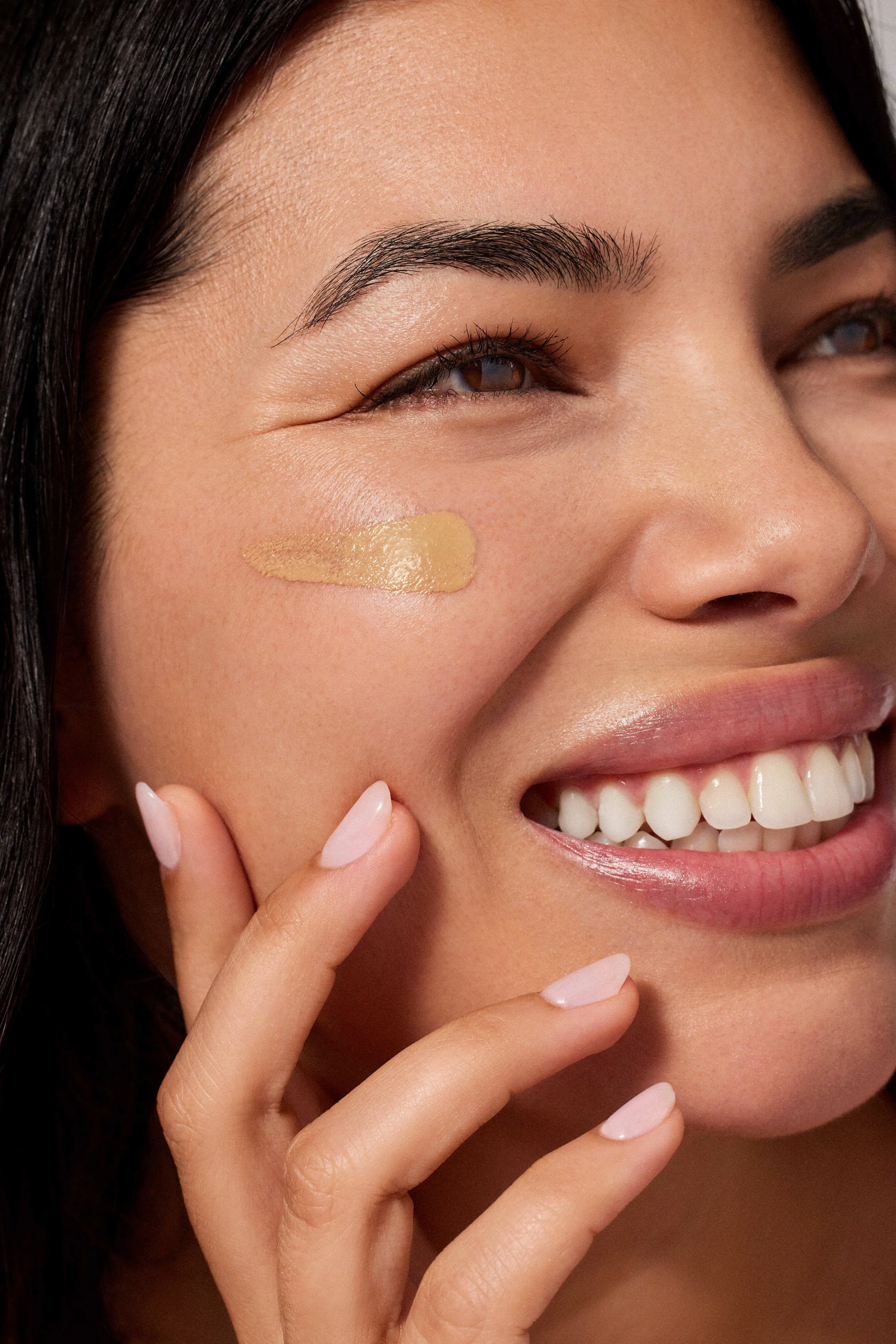Close-up of a smiling woman's face with a dollop of light yellow concealer or foundation under her eye, showing her smooth skin, dark eyebrows, and white teeth.