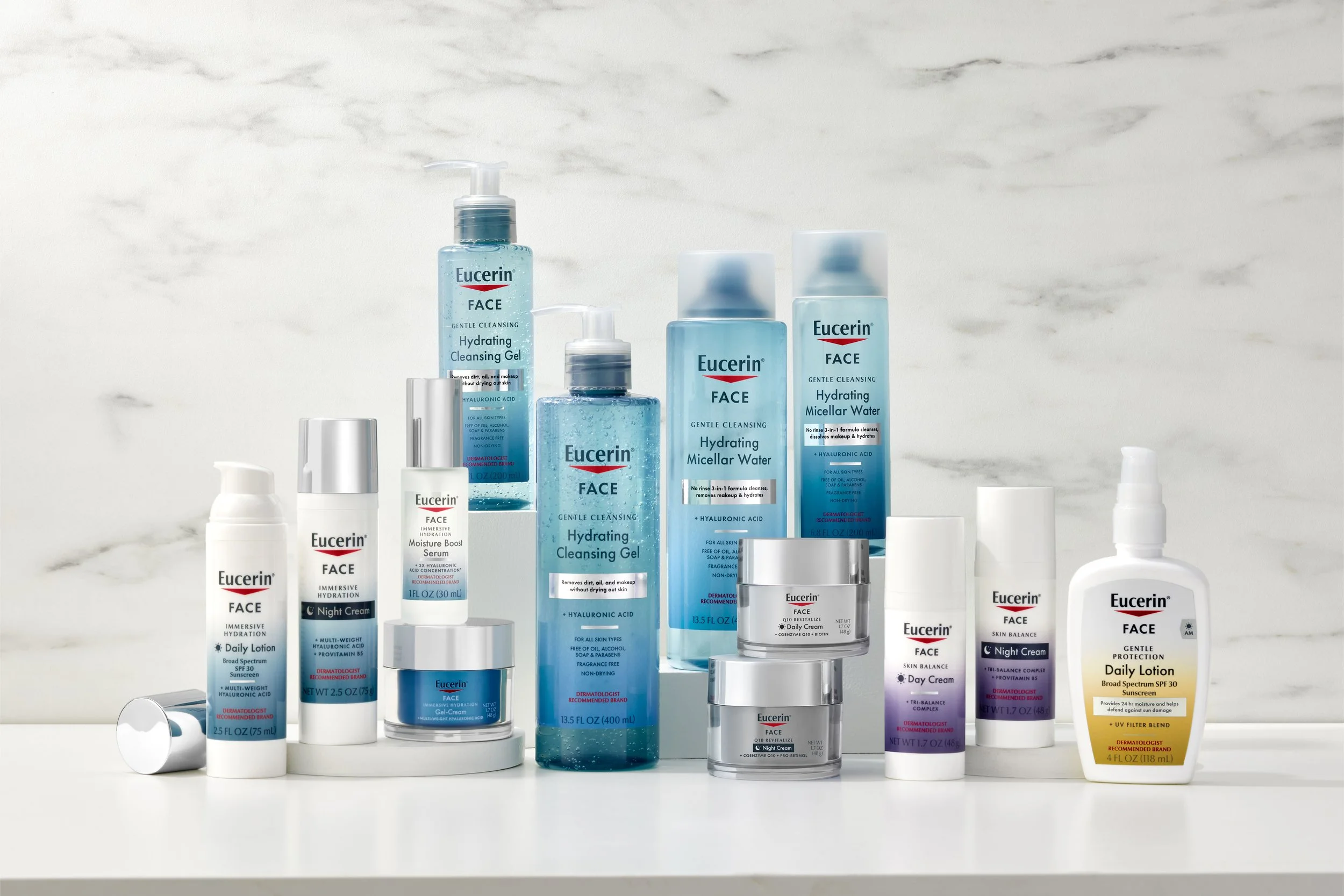 Collection of Eucerin skincare products including hydrating cleansing gel, micellar water, night cream, day cream, broad-spectrum SPF moisturizer, and various serums and lotions arranged on a white surface against a light marble wall background.