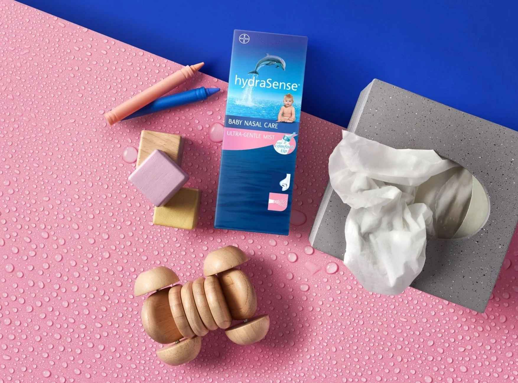 Pink textured surface with water droplets, a wooden baby toy, pastel-colored blocks, a package of HydraSense baby nasal care mist, a tissue box with tissues, and a blue background in the top corner.