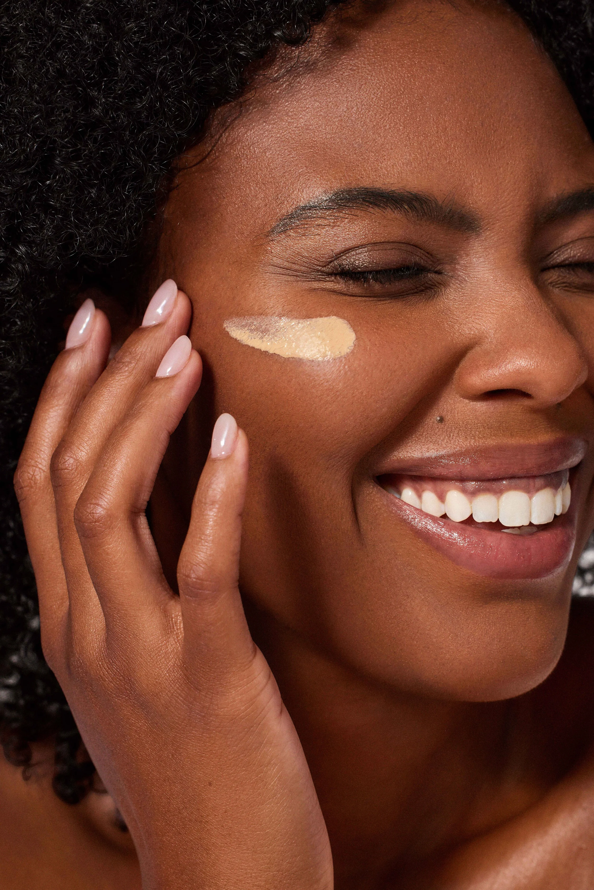 A smiling woman with curly hair touching her cheek with one hand, with a swatch of foundation on her face.