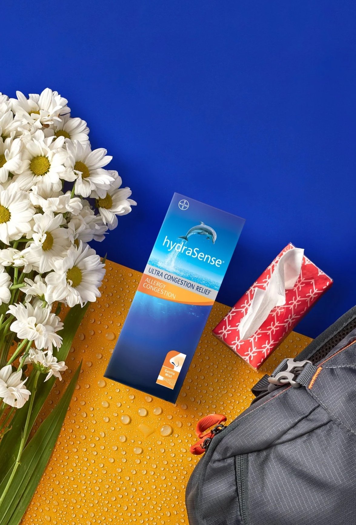 A bouquet of white daisies, a box of Hydrasense allergy congestion relief spray, a box of tissues with a red and white pattern, and part of a gray backpack on an orange surface with water droplets, against a blue background.