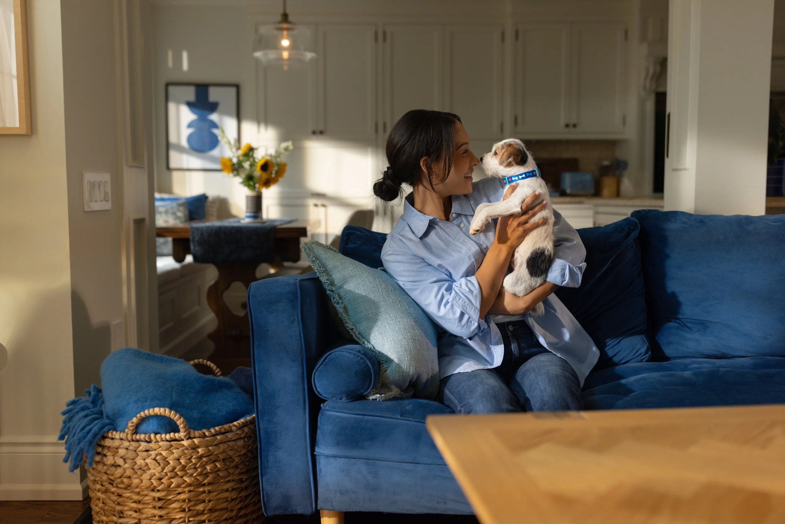 A woman with dark hair in a bun holding a small dog with a blue collar, sitting on a blue sofa in a cozy living room with kitchen in the background.
