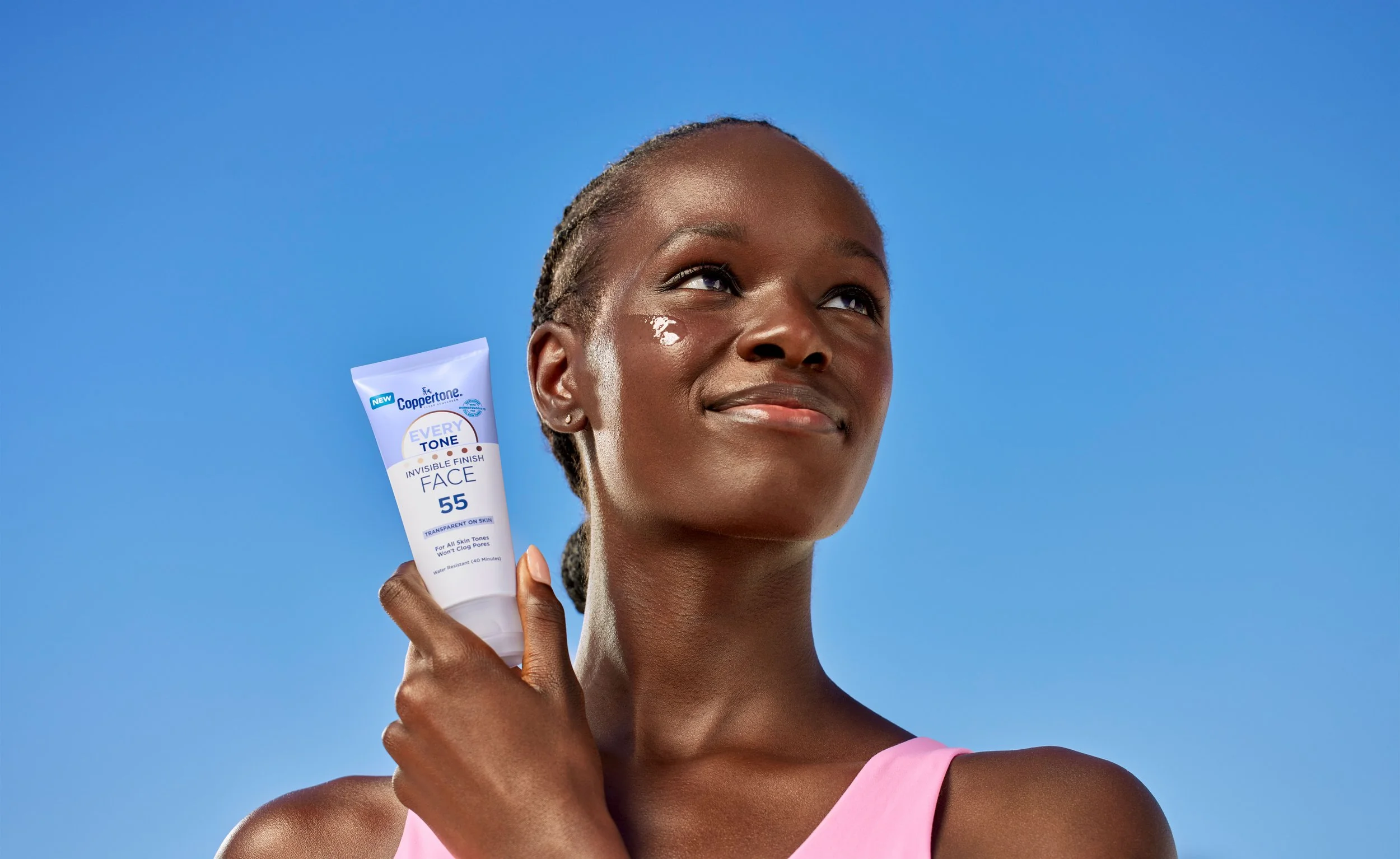 A woman with dark skin and braids is holding a tube of Coppertone sunscreen near her face. She is smiling slightly and wearing a pink top against a clear blue sky background.