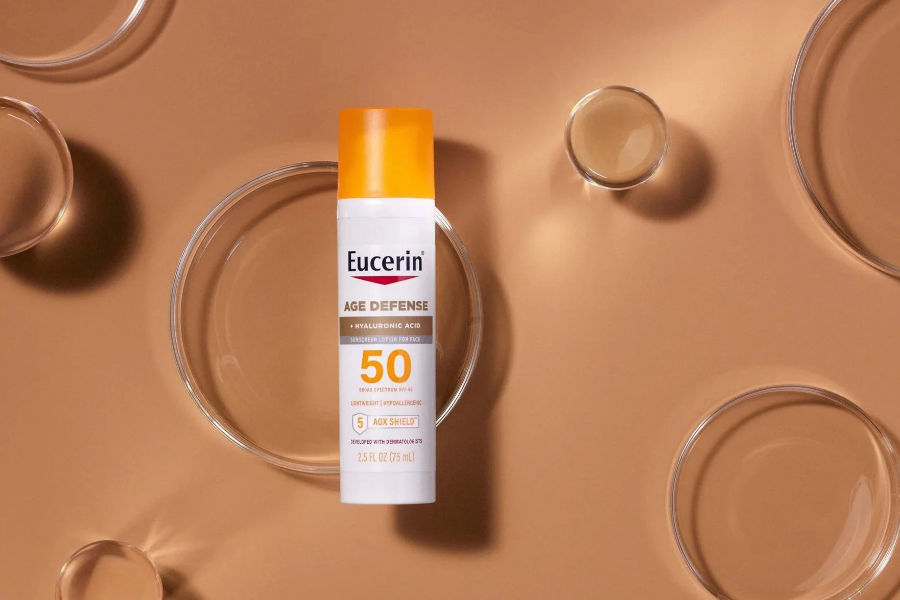 A bottle of Eucerin Age Defense SPF 50 sunscreen lotion positioned on a beige surface with several clear circular containers surrounding it.