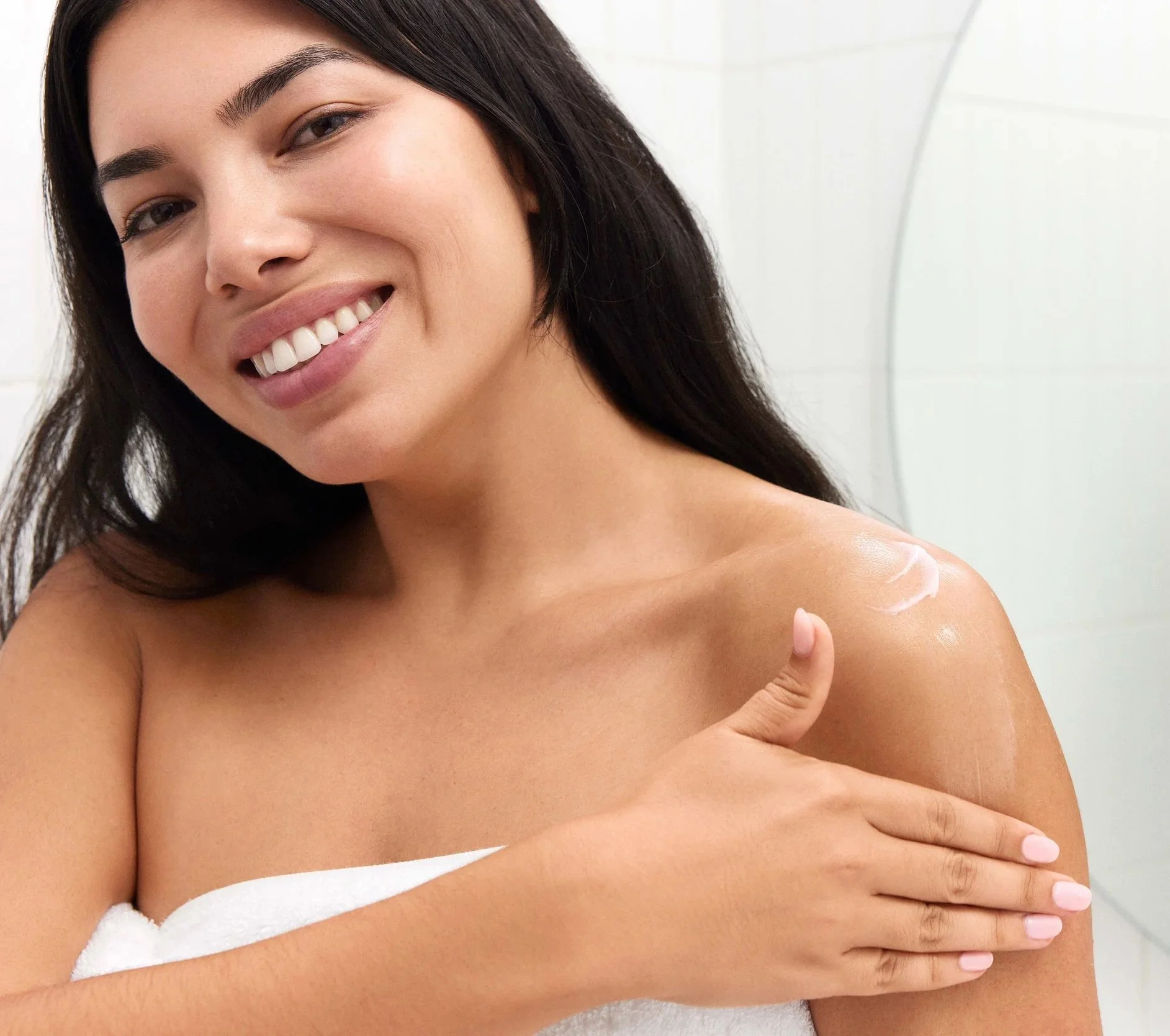 Close-up of a smiling woman with long dark hair, partially covered by a white towel, holding her shoulder with one hand, in a bright bathroom setting.