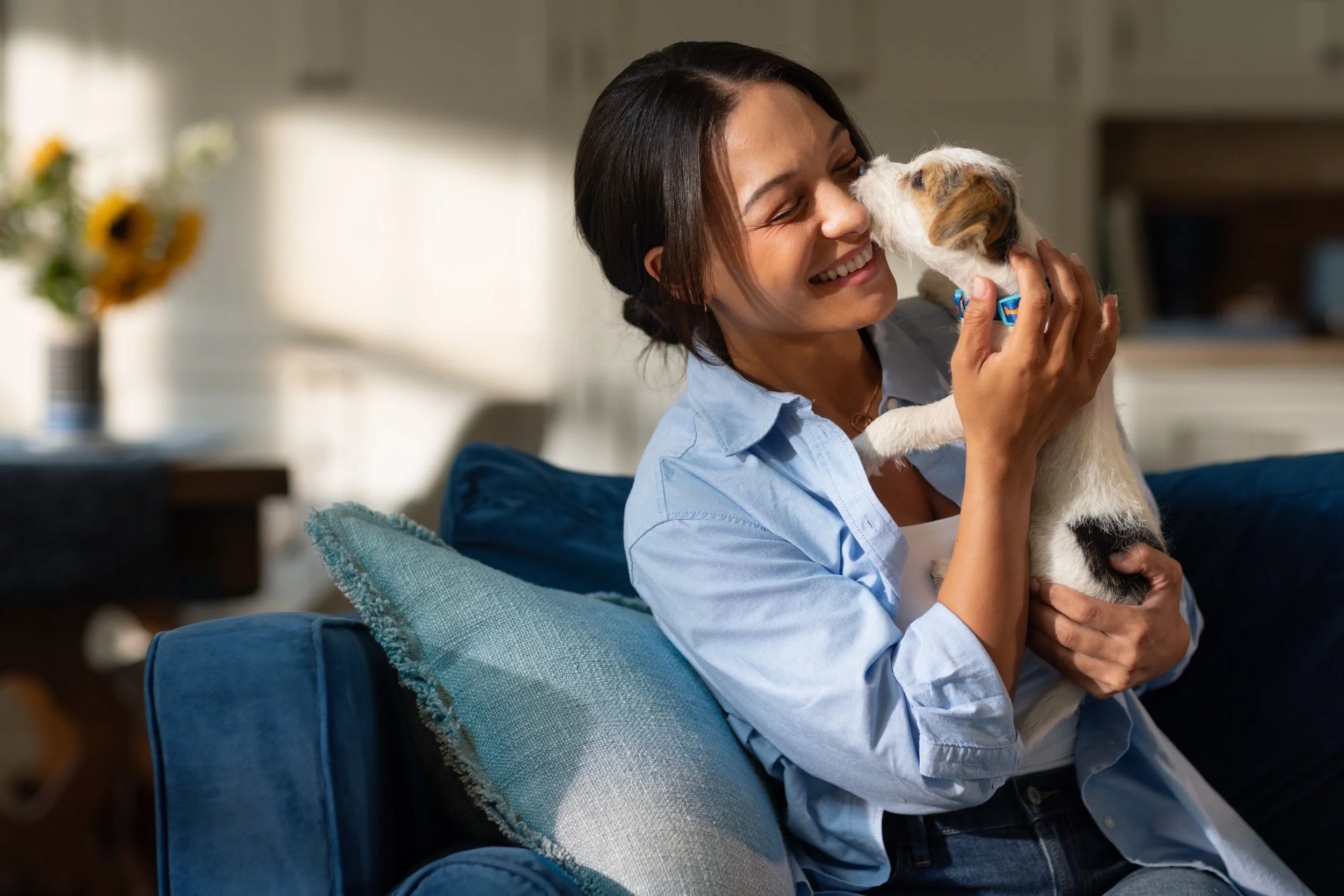 A woman with dark hair in a bun smiling and cuddling a small dog inside a cozy home. The woman is seated on a blue armchair with a light blue pillow.