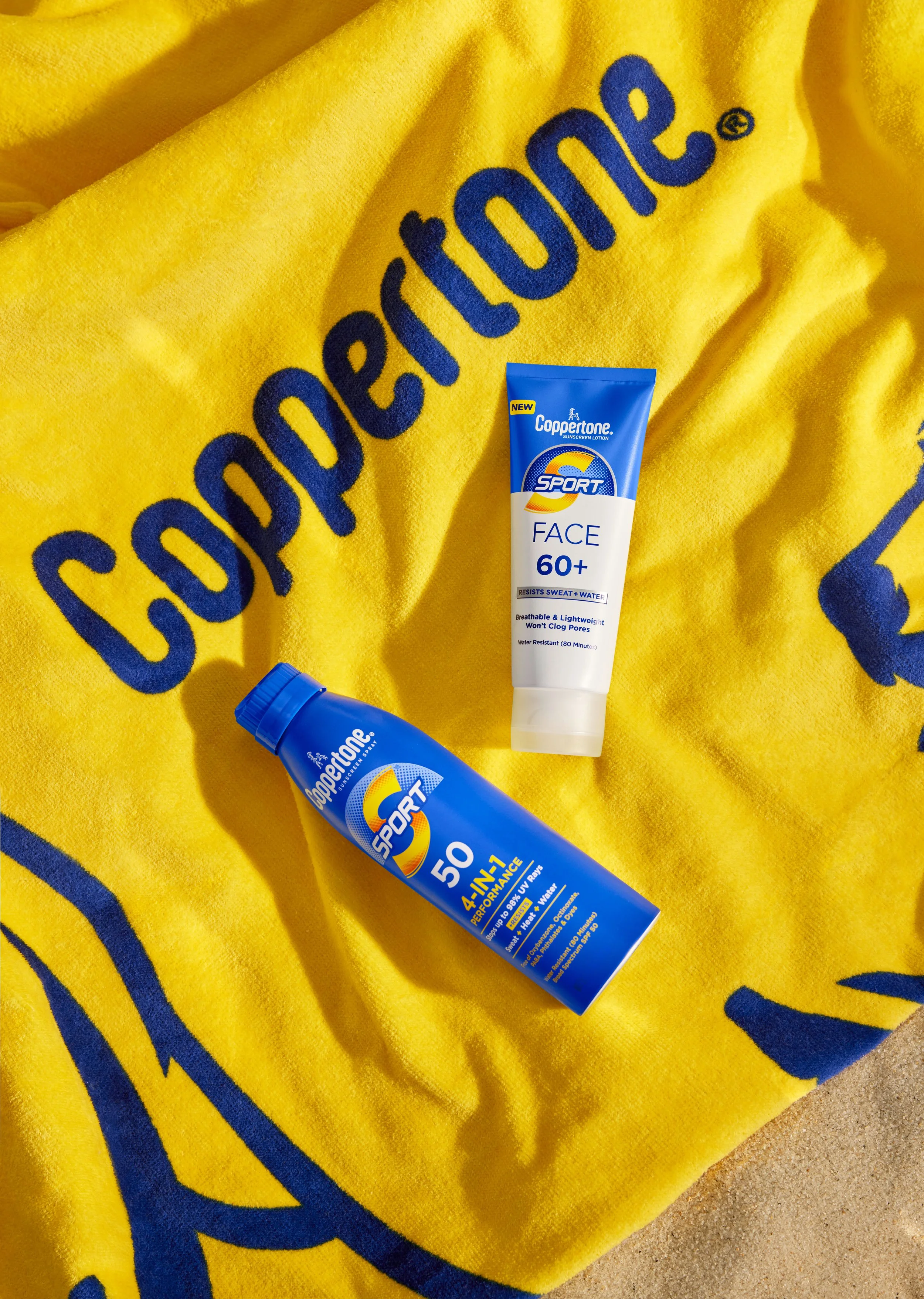 A yellow towel with the Coca-Cola logo in blue, and two bottles of Coppertone sunscreen placed on it. One sunscreen bottle is labeled "Sport Face 60+" and the other "Sport 50".