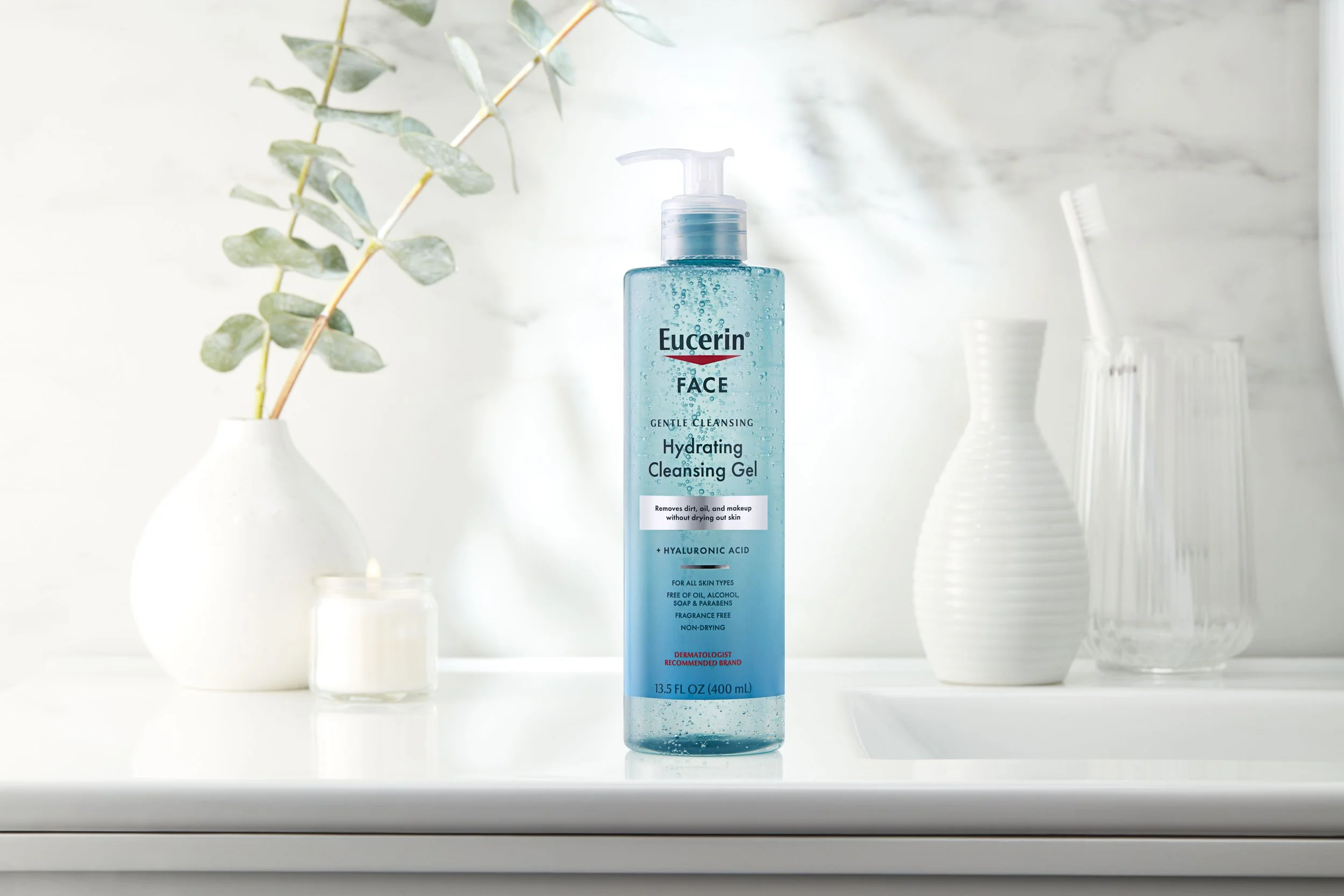 A bottle of Eucerin face hydrating cleansing gel on a white surface, with a white vase containing a green leafy branch and two white vases in the background.