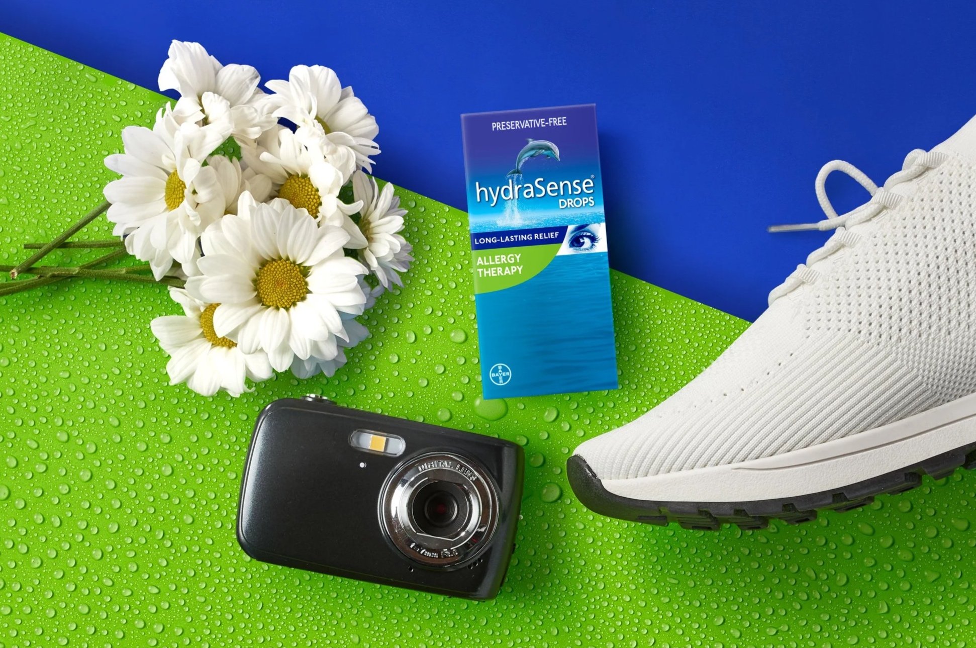 Photo of a green surface with water droplets, a bouquet of white daisies, a box of hydraSense allergy therapy drops, a white sneaker, and a black camera.