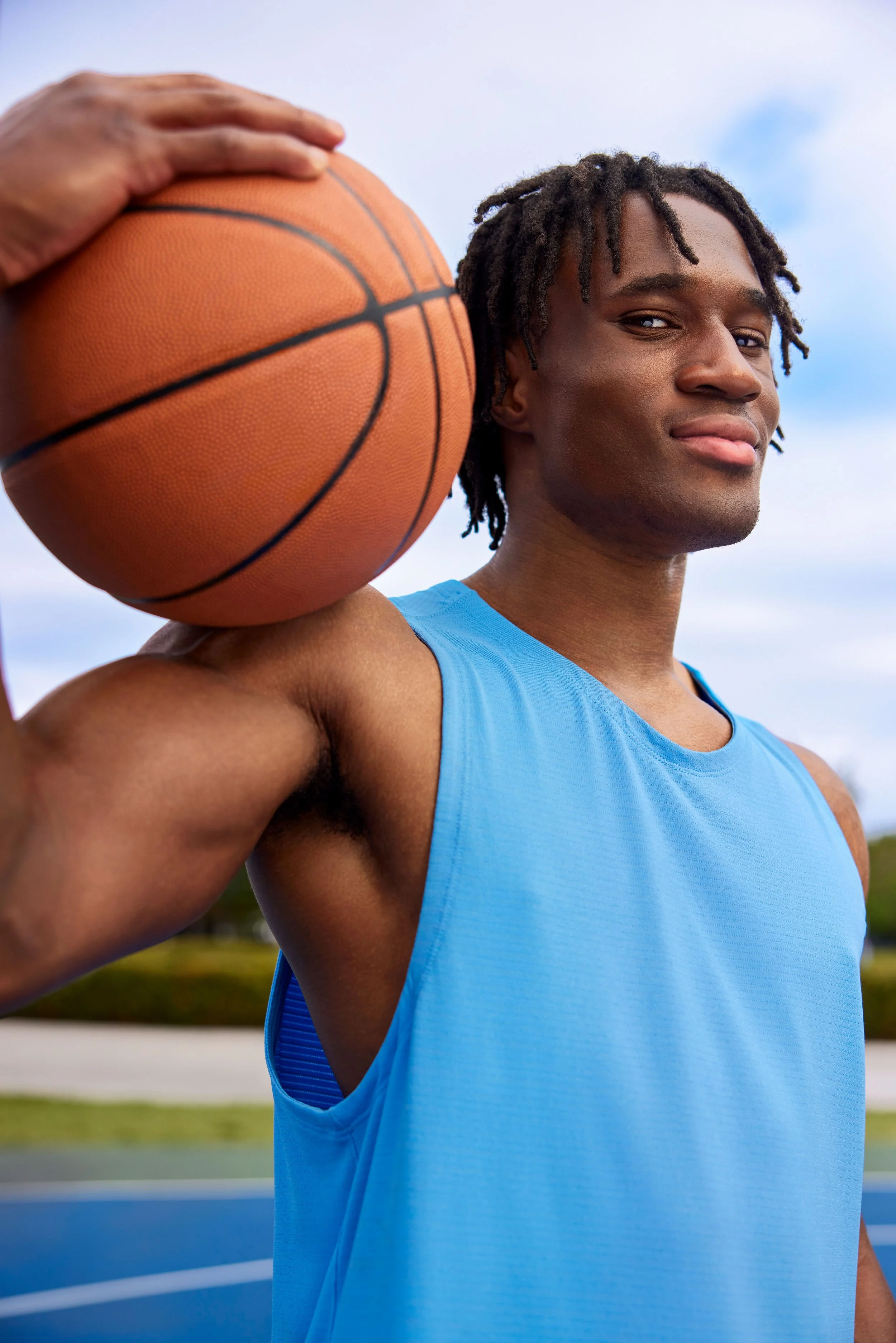 A young man outdoors holding a basketball on his shoulder, wearing a blue athletic tank top, with a clear sky in the background.