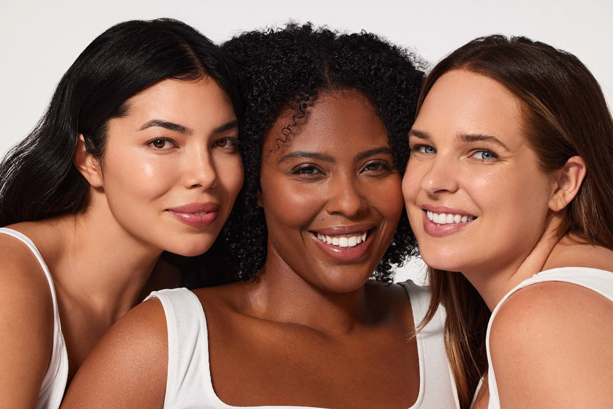 Close-up of three women smiling, with close faces, diverse ethnicities, and natural hair, in a studio setting with a white background.