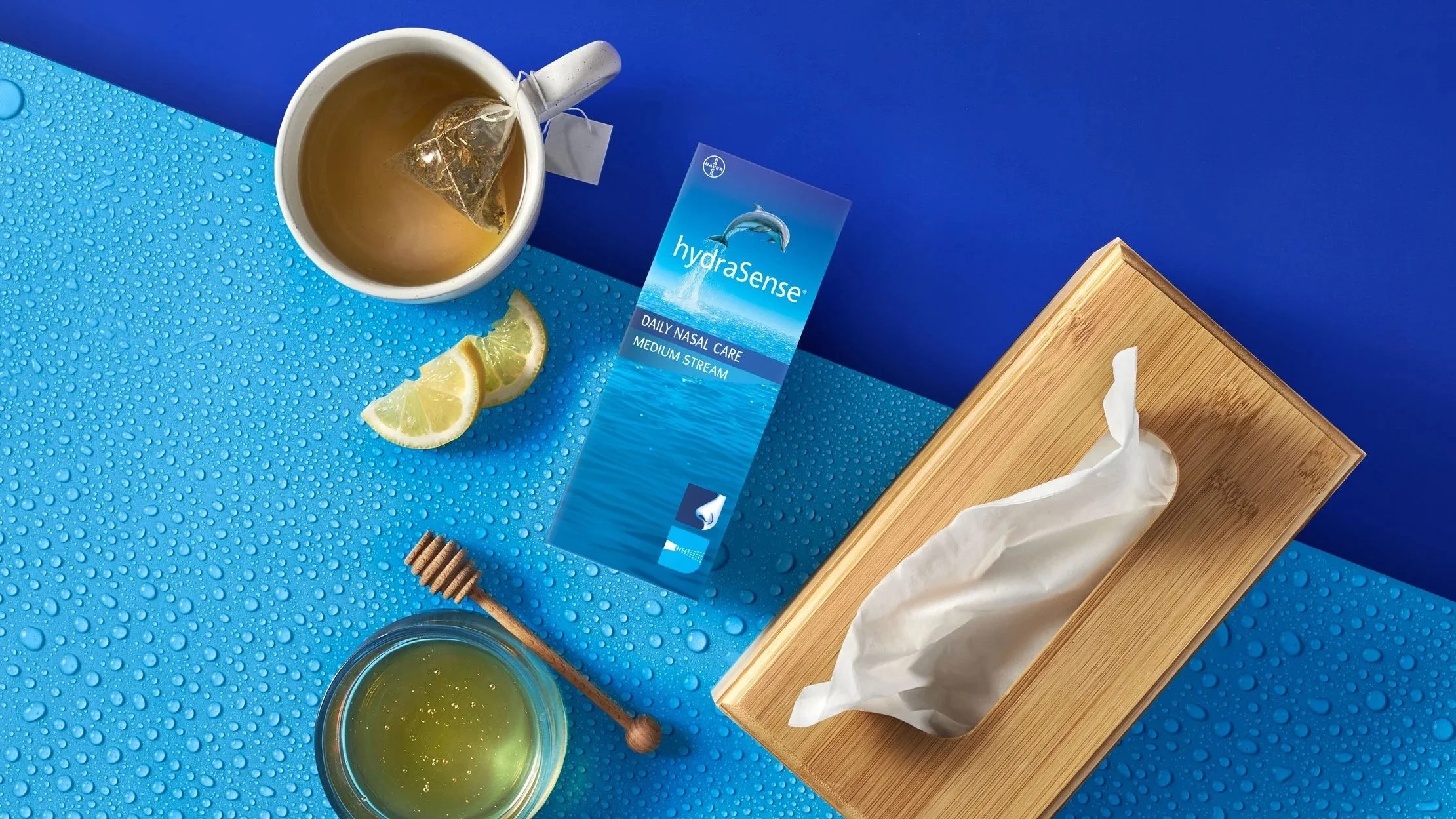 A mug of tea with a tea bag inside, lemon slices, a box of hydrasure nasal care tissues, a honey dipper, and a glass of honey on a blue, water-droplet textured surface, with a wooden box in the background.