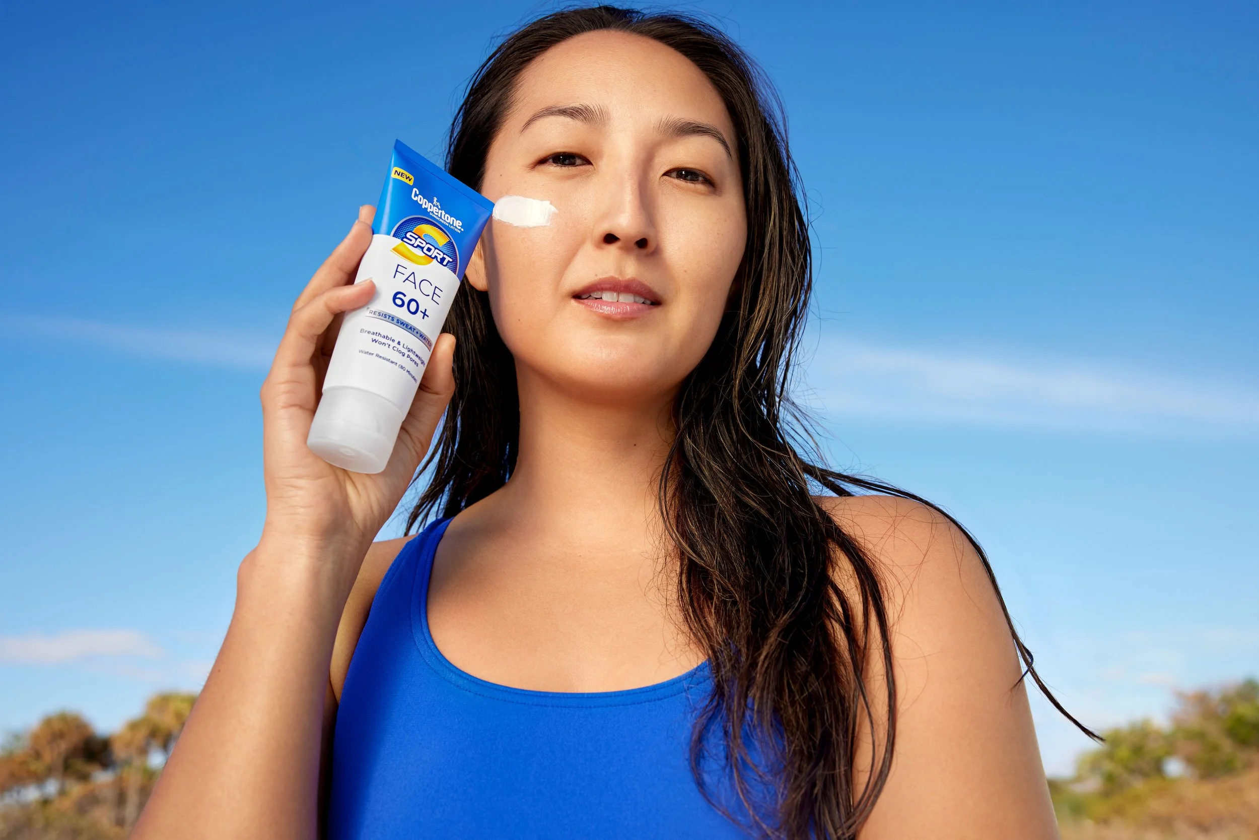 Woman holding a tube of Coppertone sunscreen near her face outdoors against a blue sky.