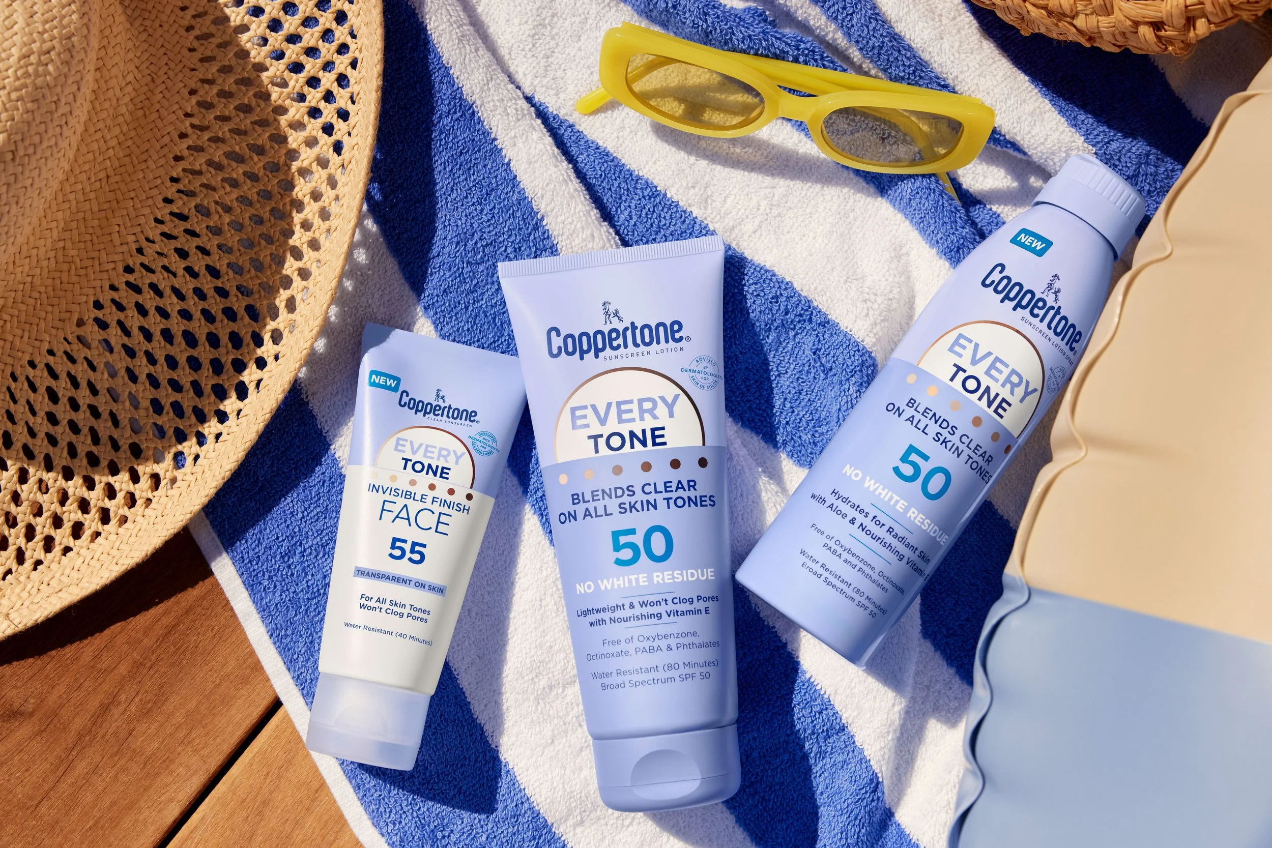 A collection of Coppertone sunscreen products, including lotions and sprays, laid out on a blue and white striped towel with a woven straw hat and yellow sunglasses nearby.