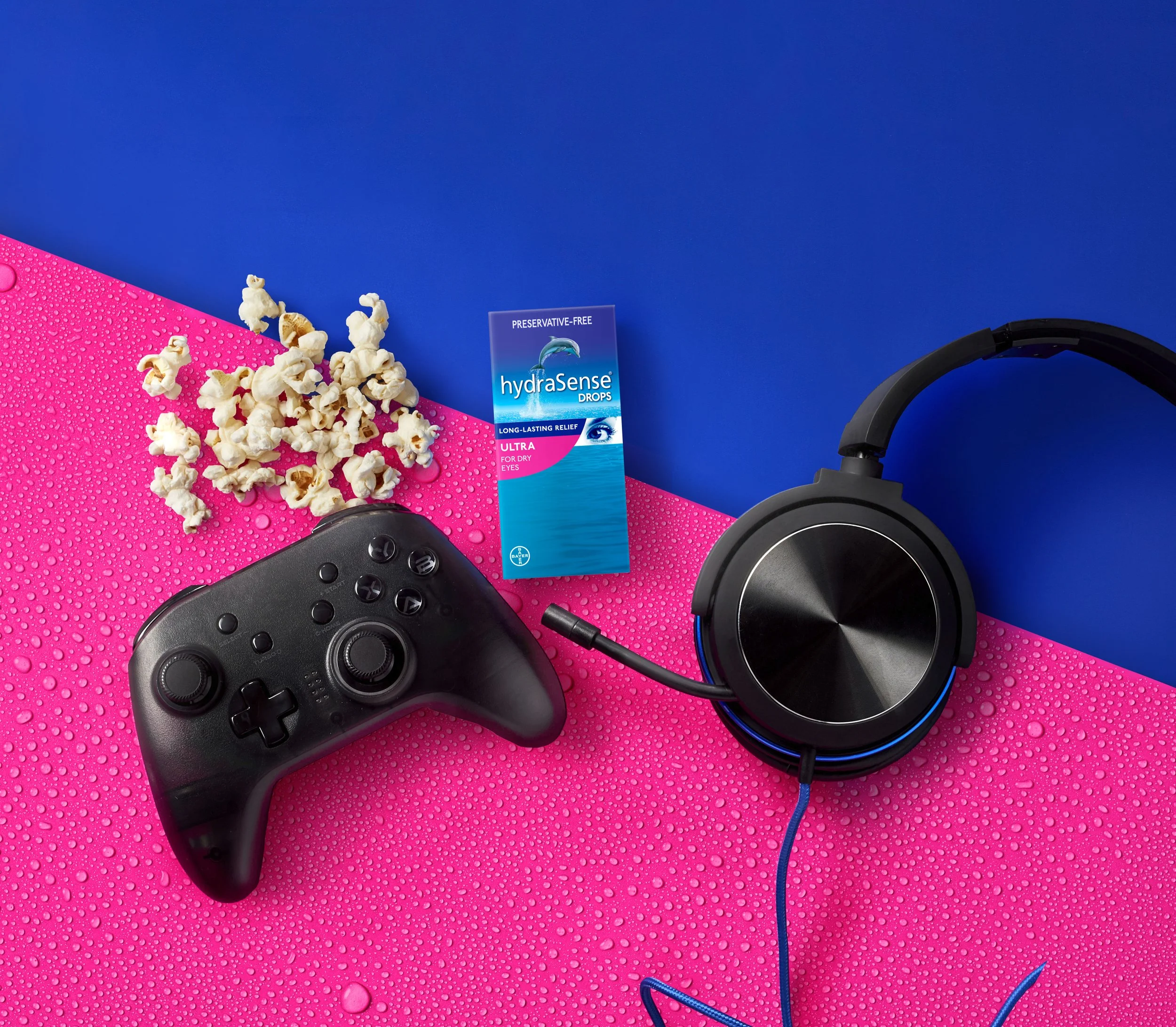 Popcorn spilled on a pink surface with water droplets, a pair of black headphones, a black game controller, and a box of HydraSense eye drops on a blue background.