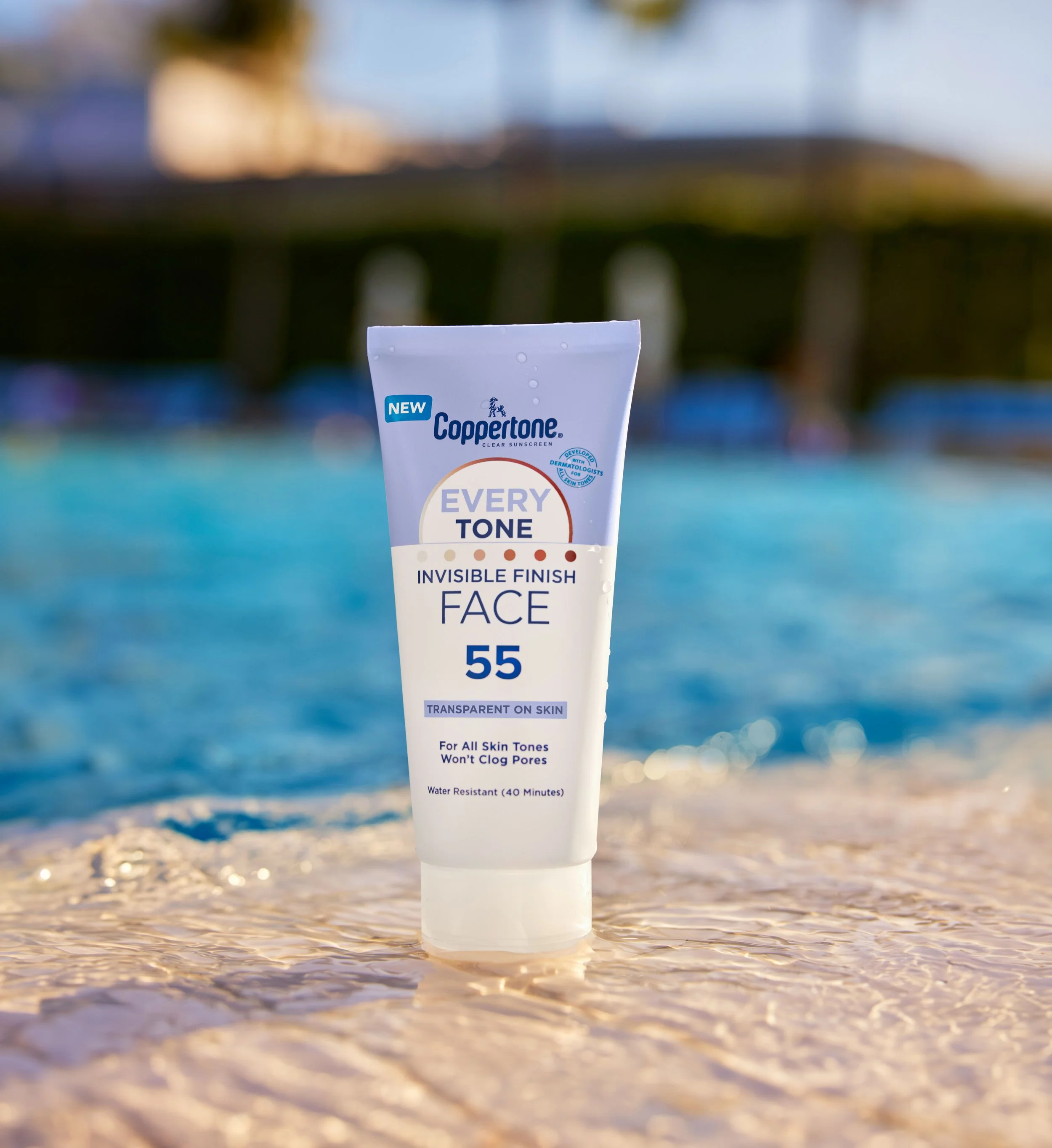 A tube of Coppertone face sunscreen labeled 'Every Tone 55' partially submerged in shallow water at the beach, with a blurred background of the ocean and sky.