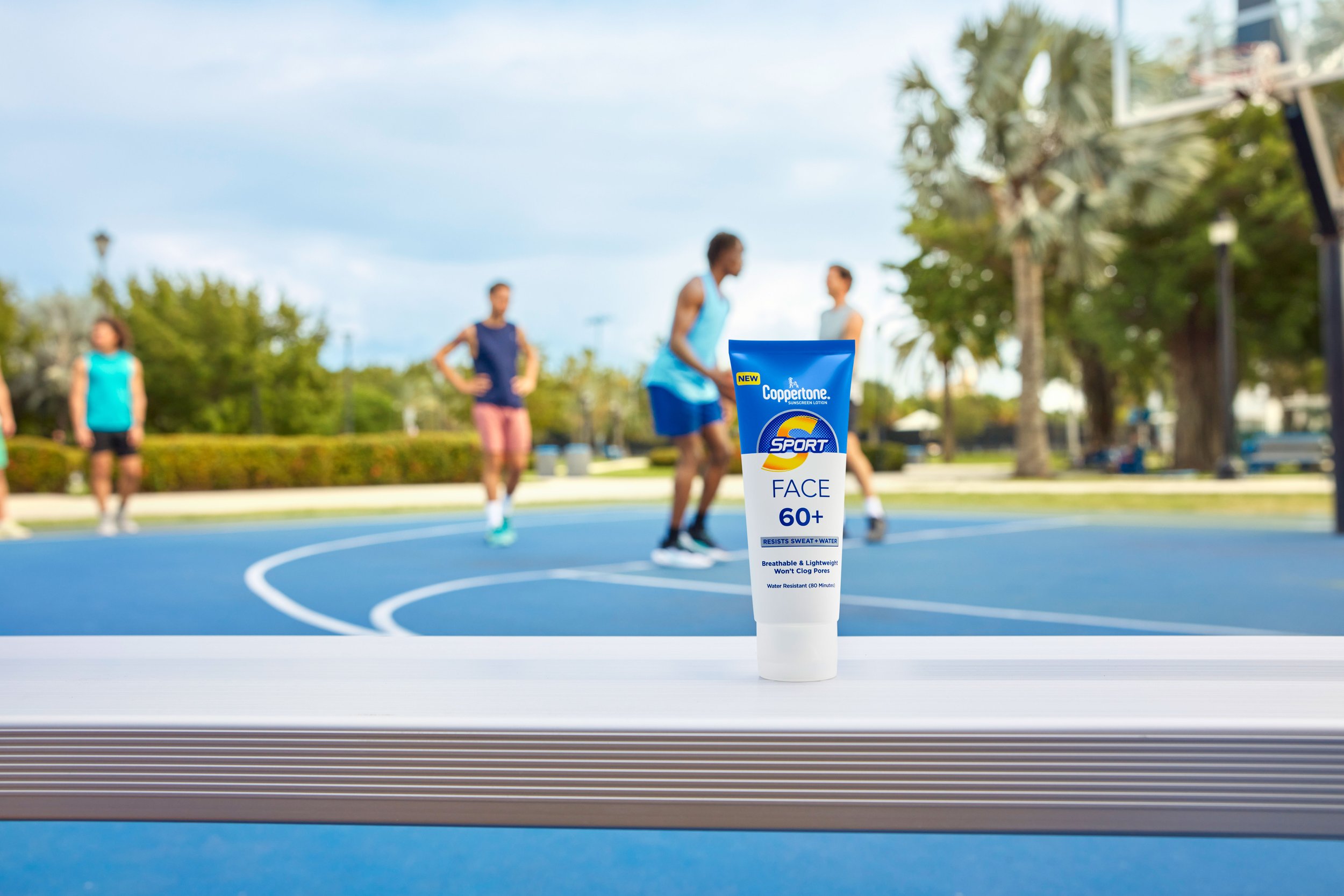 Tube of Coppertone sunscreen lotion labeled 'FACE 60+', placed on a table at an outdoor basketball court. In the background, a group of people is playing basketball on the court with trees and a clear sky around.