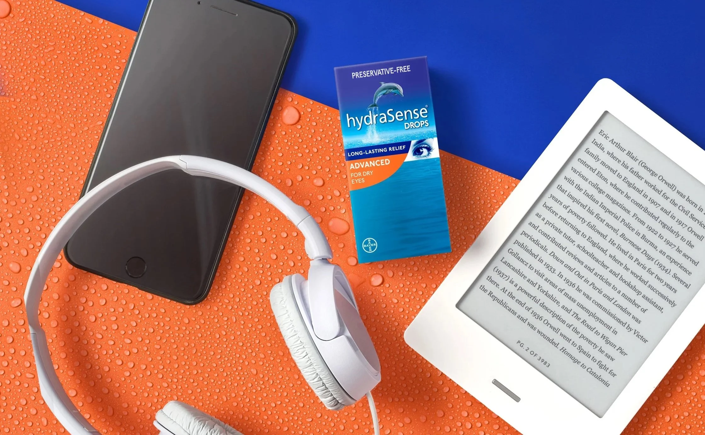 A flat lay of a smartphone, white headphones, a box of hydraSense nasal spray, an e-reader displaying text, all placed on a surface with orange and blue backgrounds, and water droplets on the orange part.