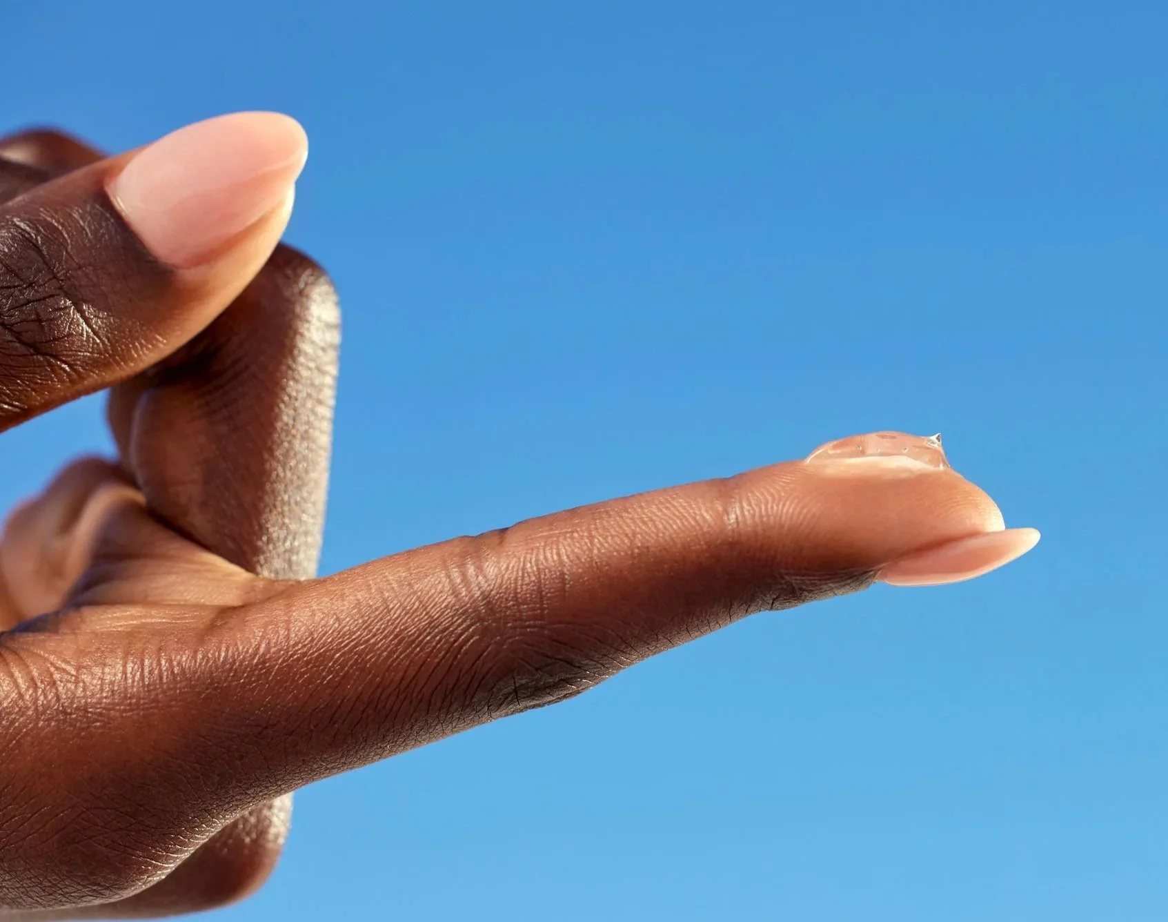 Close-up of a hand with dark skin holding a small, transparent water droplet between the thumb and index finger against a clear blue sky.