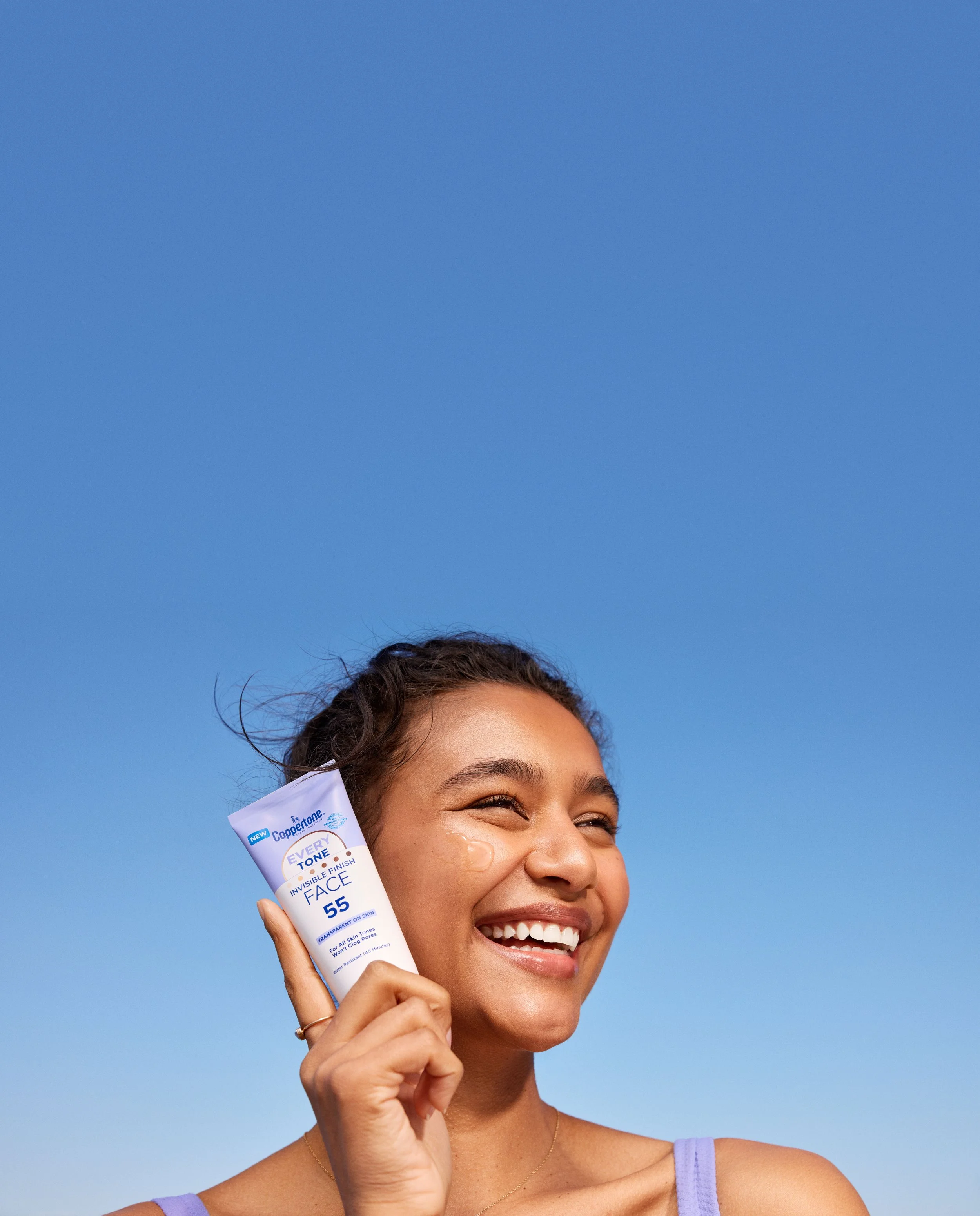 A young woman with a radiant smile holding a tube of facial sunscreen against a clear blue sky.