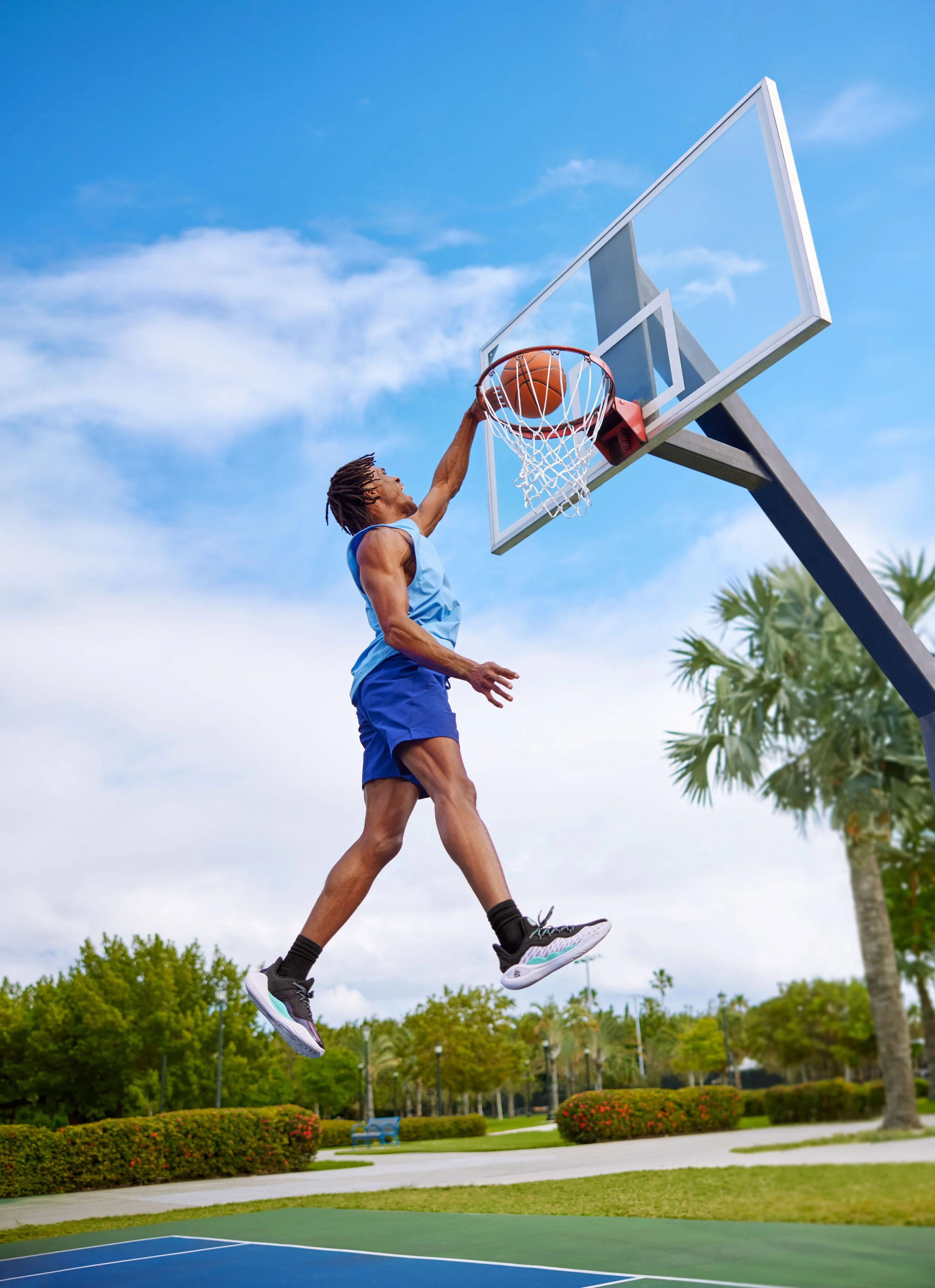 A man is mid-air jumping to dunk a basketball into the hoop on an outdoor court, with trees and blue sky in the background.