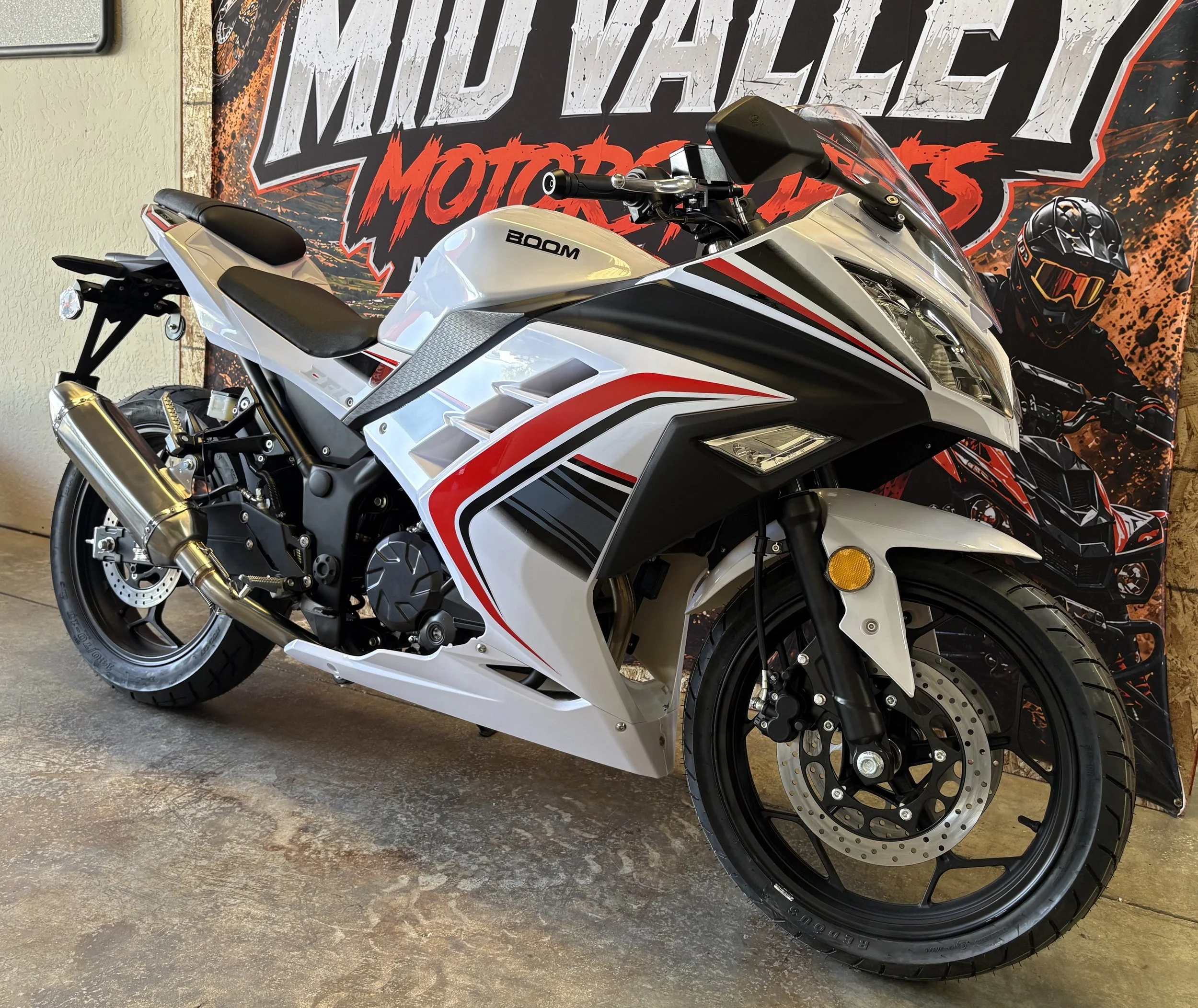 A white and red Honda CBR motorcycle displayed indoors in front of a large promotional poster with the text 'MOTO LIFE MOUNTAIN' and an image of a motorcyclist in black gear and helmet.