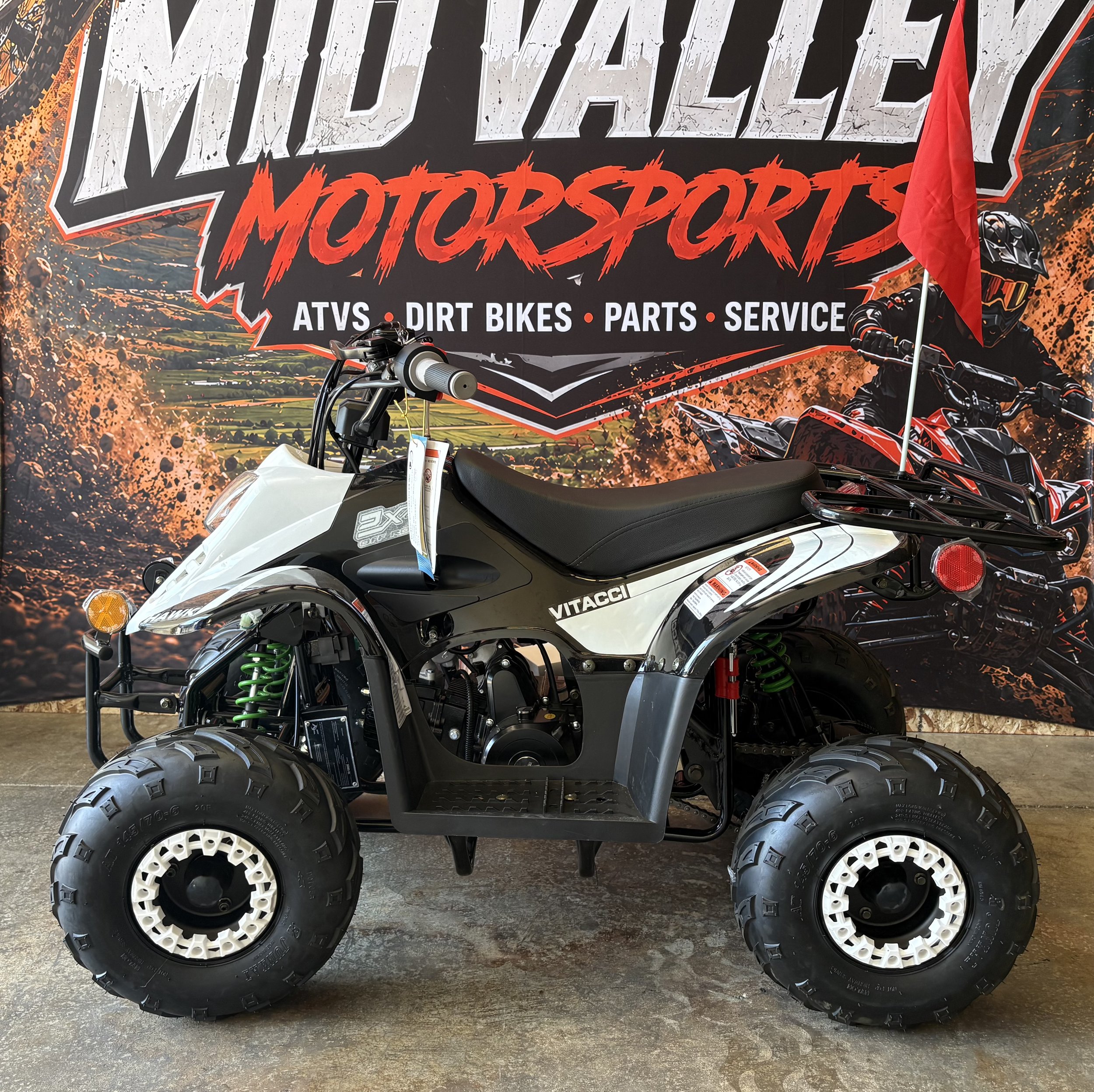 Off-road all-terrain vehicle parked in front of a motocross dealership banner that displays ATV, dirt bikes, parts, and service, with a red flag attached to the vehicle.