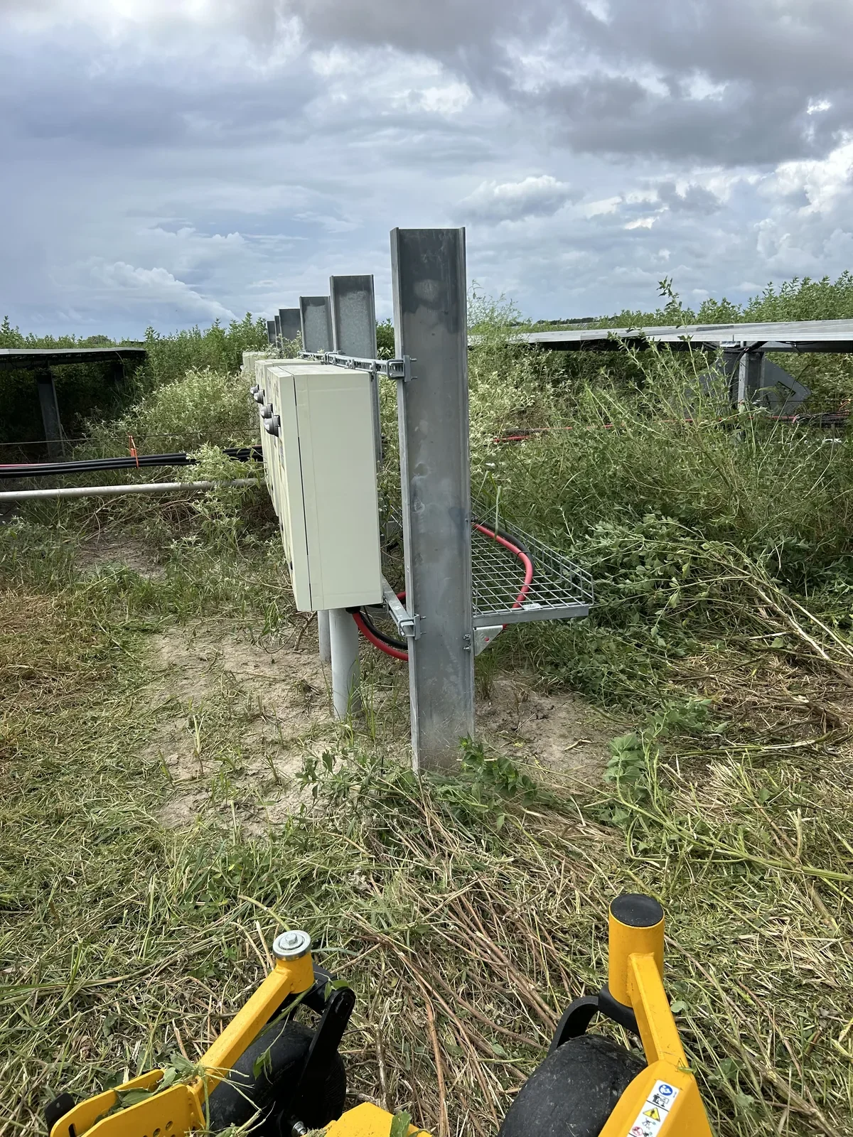 Electrical equipment mounted on metal posts in a grassy field with solar panels and cloudy sky in the background.