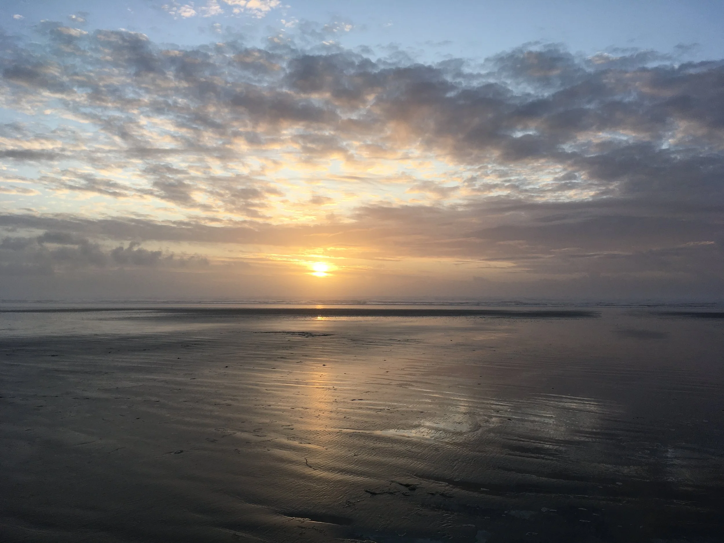 A sunset over a beach with clouds partly covering the sky. The sun is near the horizon, casting a reflection on the wet sand and gentle waves.