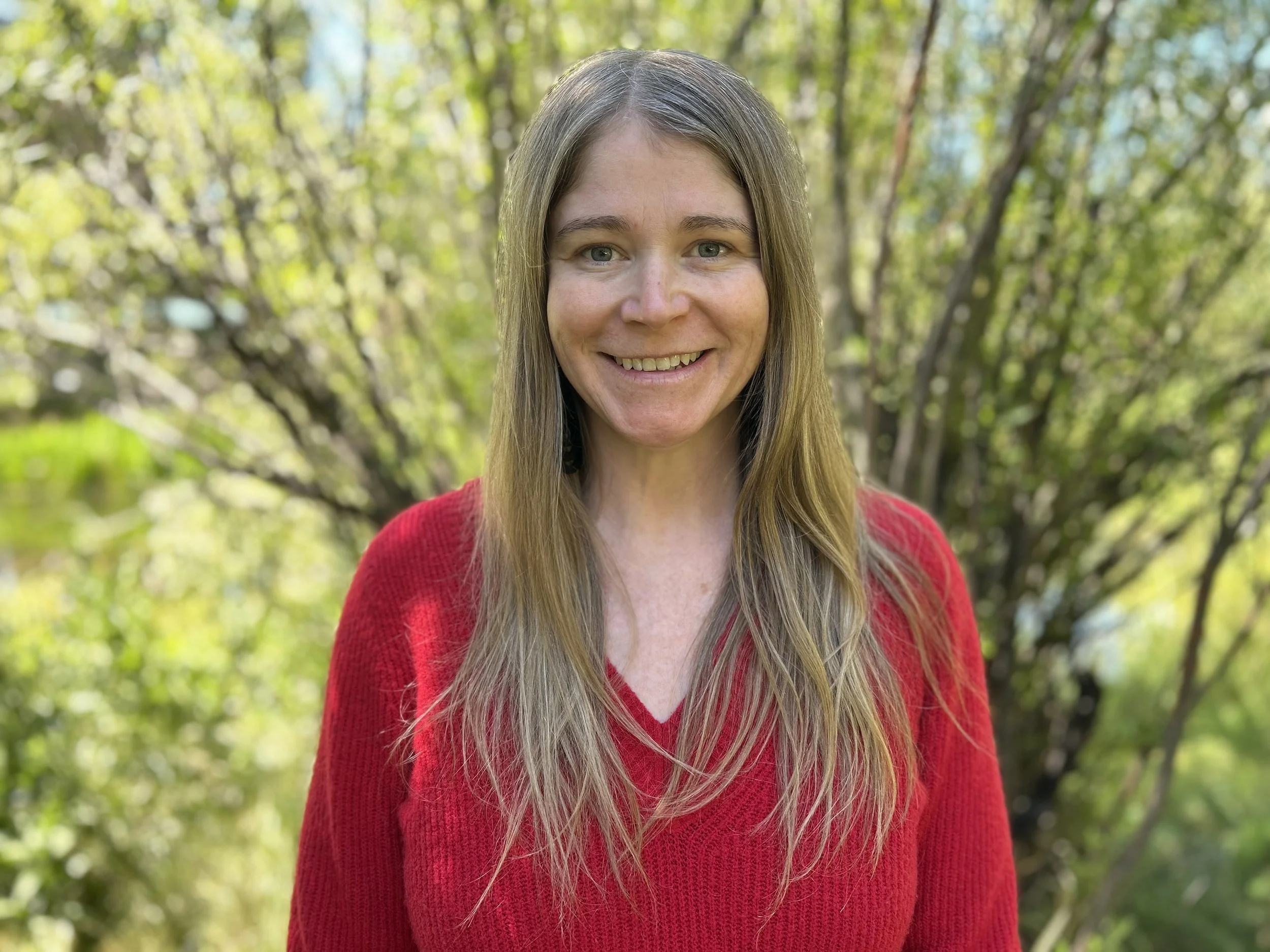A woman with long blonde hair wearing a red sweater, smiling outdoors with green trees and sunlight in the background.