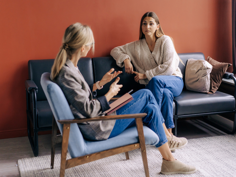 Two women having a conversation in a waiting room; one is taking notes, the other listens attentively.