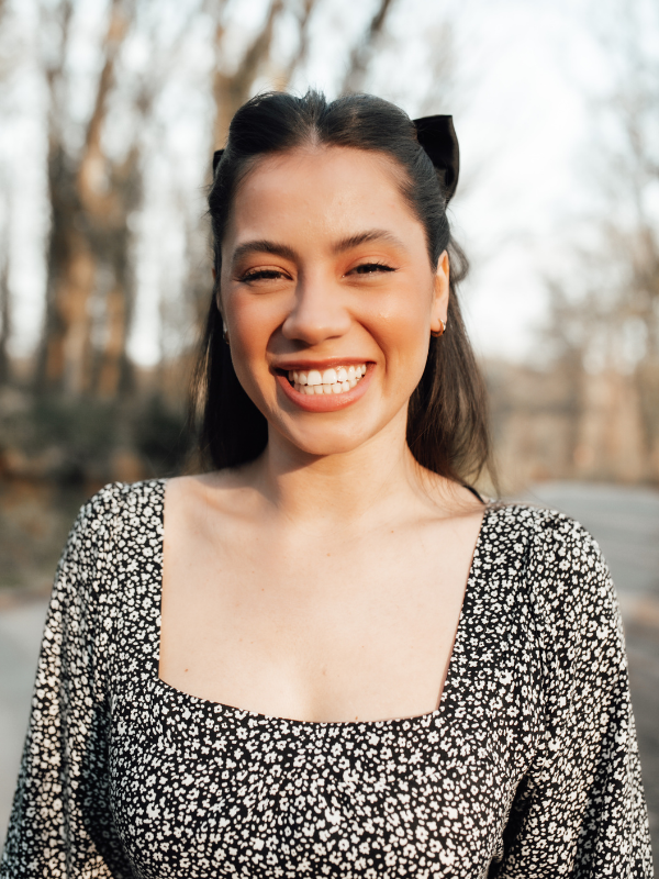 A young woman with dark hair, smiling broadly with her eyes closed, standing outdoors with blurred trees in the background.