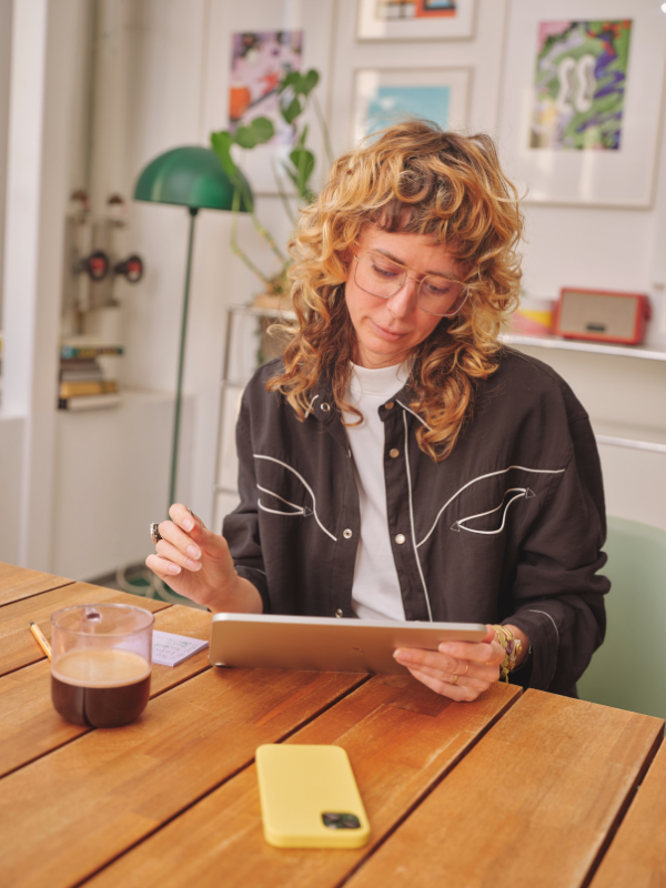 A woman with curly blonde hair, wearing glasses and a black shirt with white line details, is sitting at a wooden table, looking at a tablet. On the table, there is a yellow phone case, a cup of coffee, and a small notepad with a pen. In the background, there are framed pictures on the wall, a green desk lamp, and a potted plant.