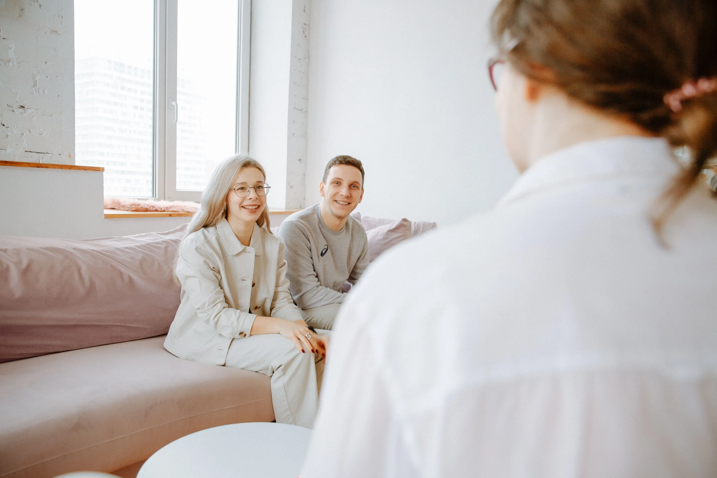 Two people sitting on a bed, smiling and talking with a third person whose back is to the camera, in a bright room with large windows.
