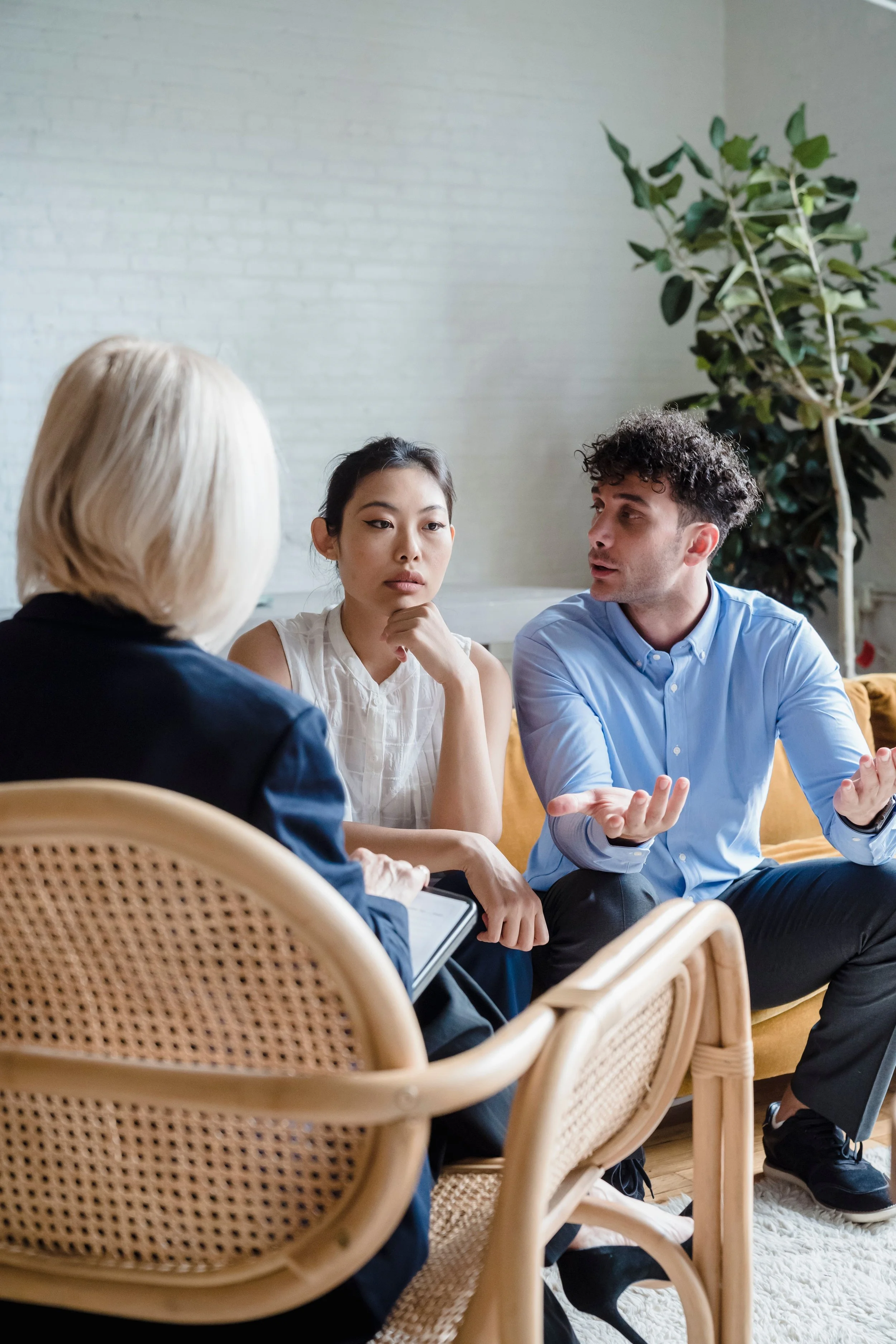 A group of four people engaged in a discussion in a bright room, with a woman in a white sleeveless shirt and a man in a blue shirt gesturing as they speak. There is a large green plant in the background.